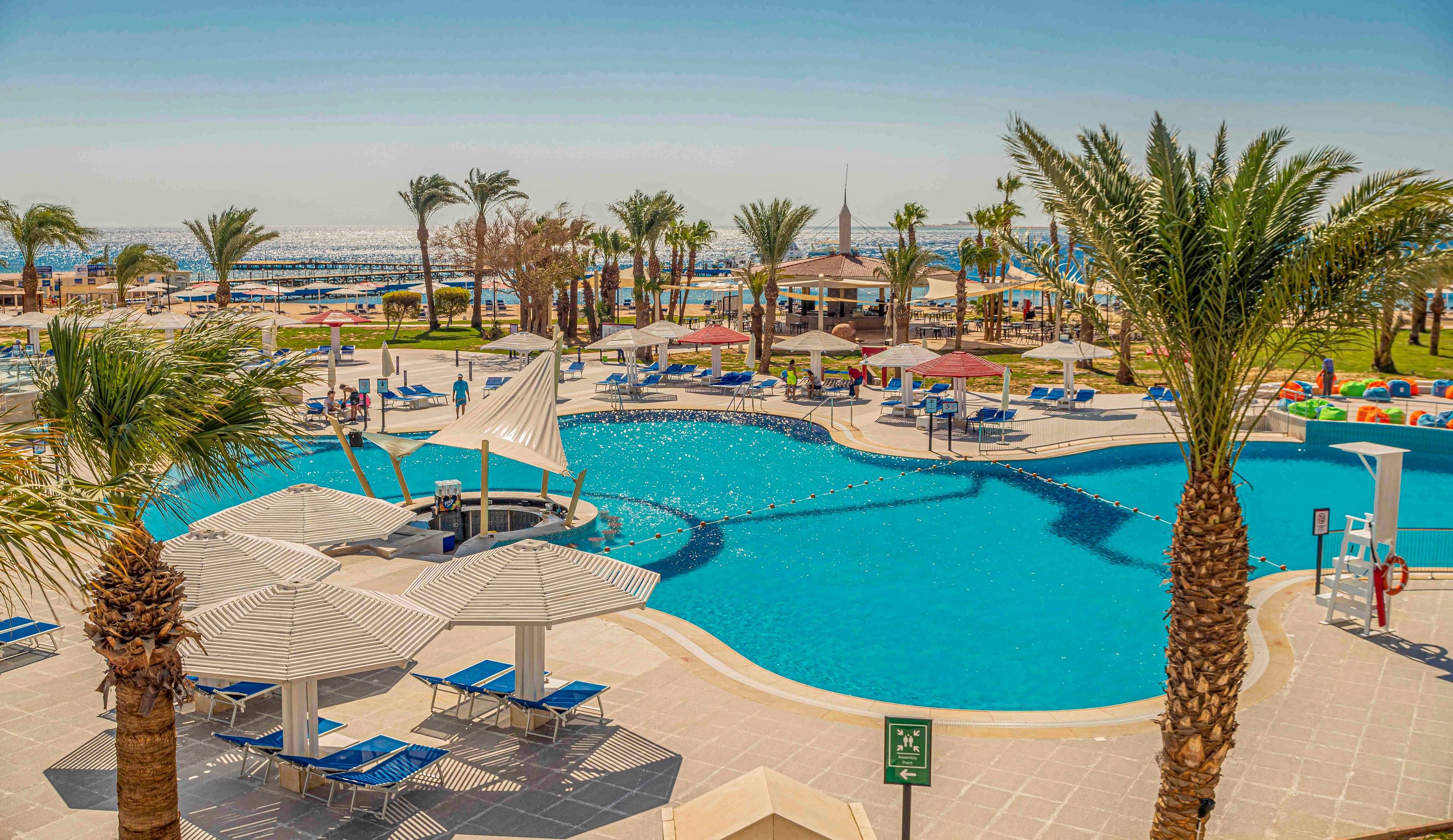 A large outdoor swimming pool is surrounded by tall palm trees and sun loungers at a seaside resort. Blue lounge chairs and umbrellas line the tiled pool deck, creating shaded relaxation areas. In the background, guests enjoy the beach and water activities near the shoreline. The bright daylight and clear sky give the scene a warm, tropical vacation atmosphere.