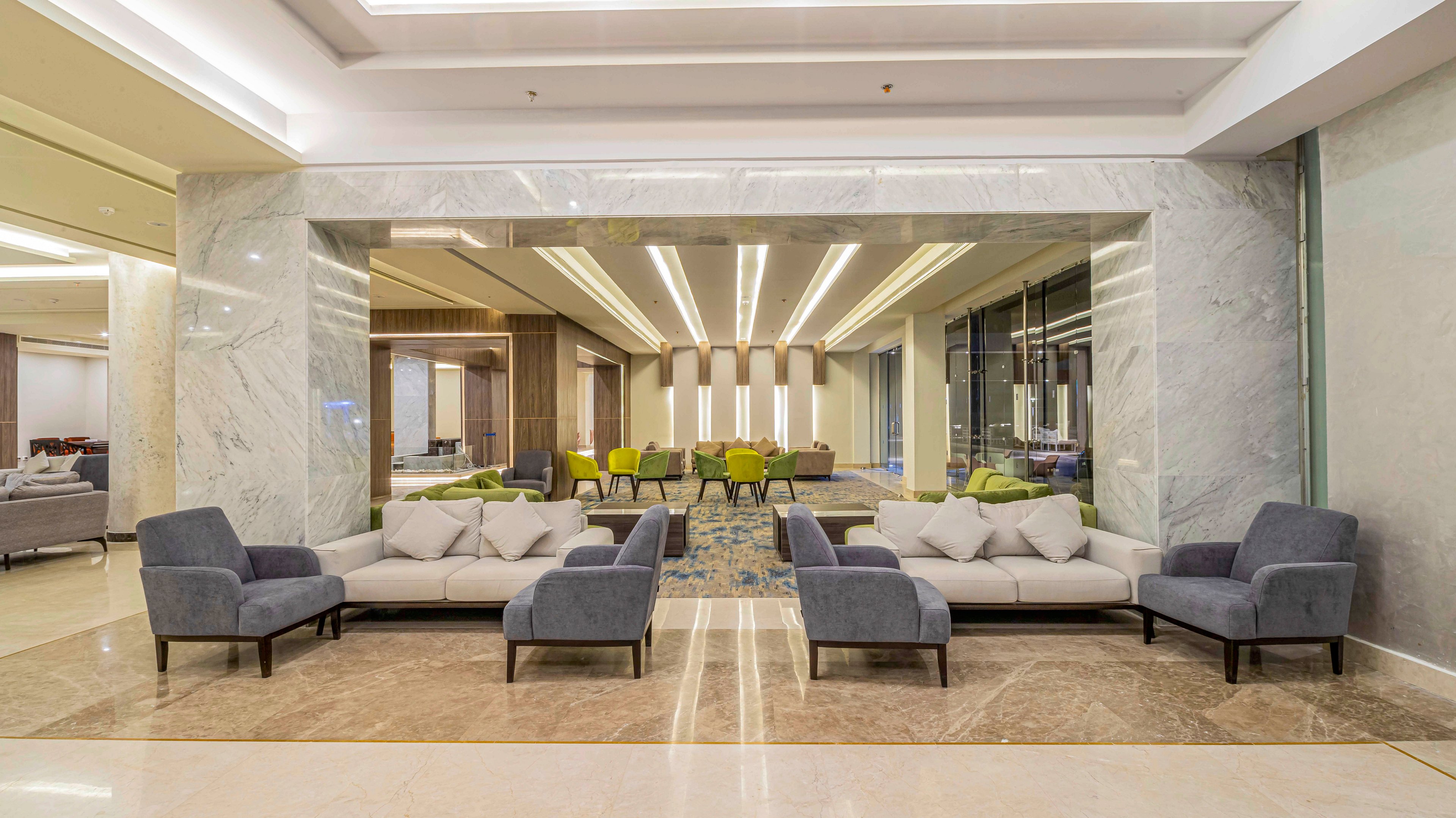 Spacious modern lobby area featuring contemporary sofas and armchairs arranged in symmetrical seating groups. The setting includes polished marble floors, large stone wall panels, and a reflective ceiling with linear recessed lighting. In the background, additional lounge chairs and tables create a comfortable waiting or meeting space. The overall design emphasizes clean lines, neutral tones, and a bright, welcoming atmosphere.