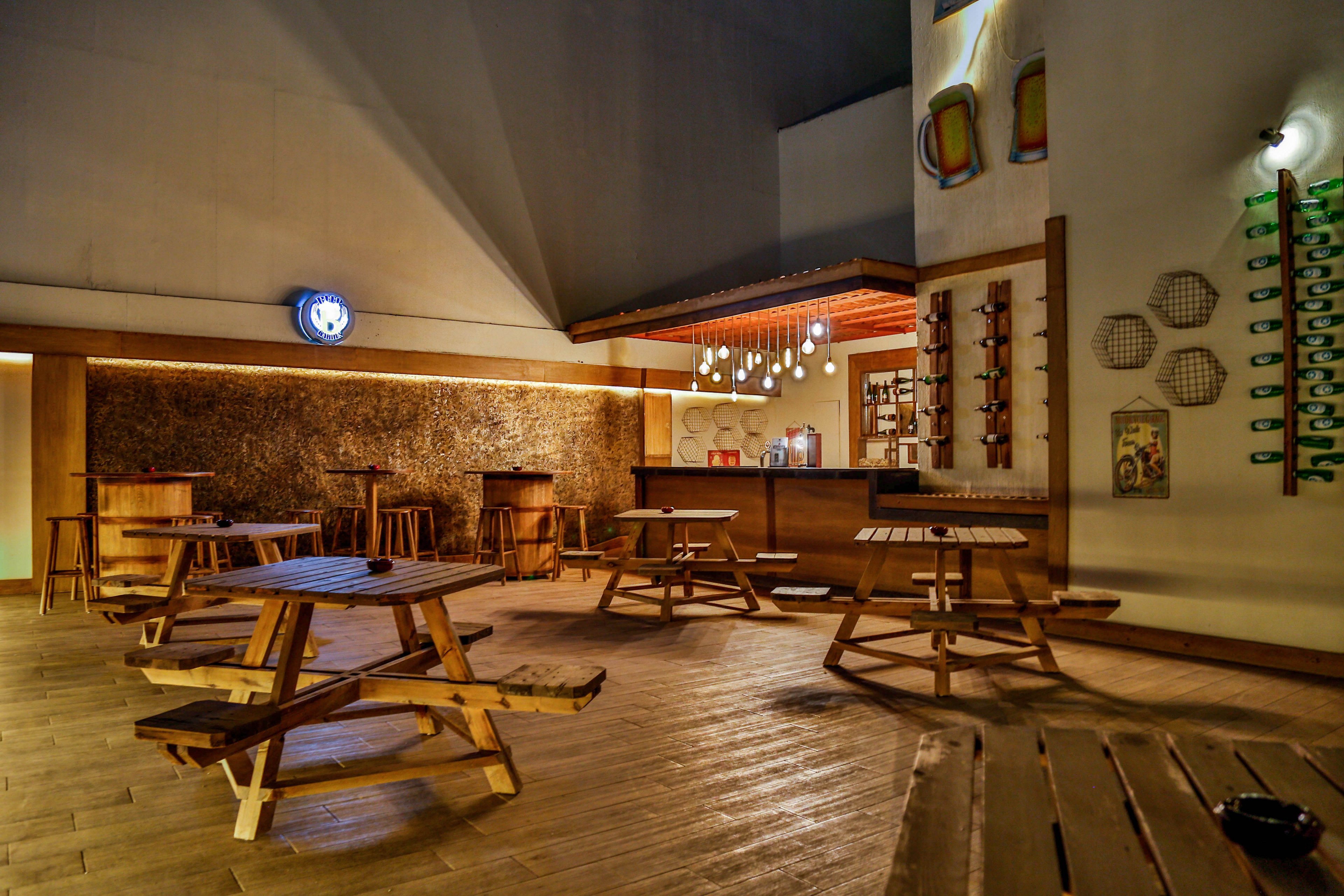 An inviting bar interior features several wooden picnic-style tables and benches arranged on a polished wooden floor. The bar counter at the back is warmly lit with hanging pendant lights and bottles displayed on shelves. Decorative elements such as wall-mounted rackets and bottle displays add character to the space. The scene is empty of people, creating a calm, relaxed atmosphere suitable for social gatherings.