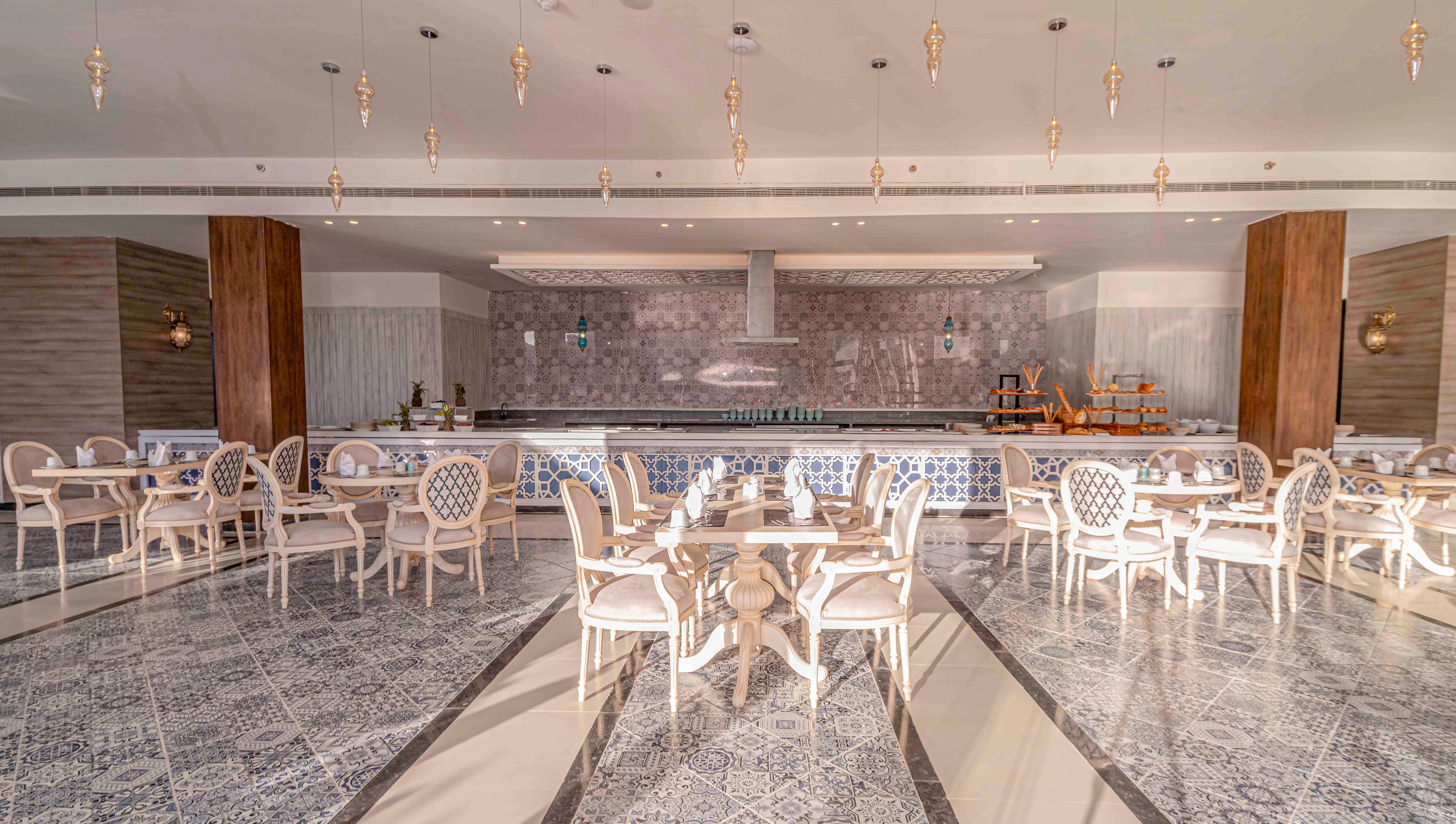 Spacious restaurant interior featuring elegant white chairs and round tables arranged in neat rows. The setting includes a long open counter or buffet area against a stone-textured wall. Decorative floor tiles with geometric patterns lead toward the serving area, complemented by warm wooden columns. Soft, warm lighting from ceiling fixtures and wall sconces creates an inviting, contemporary dining atmosphere.