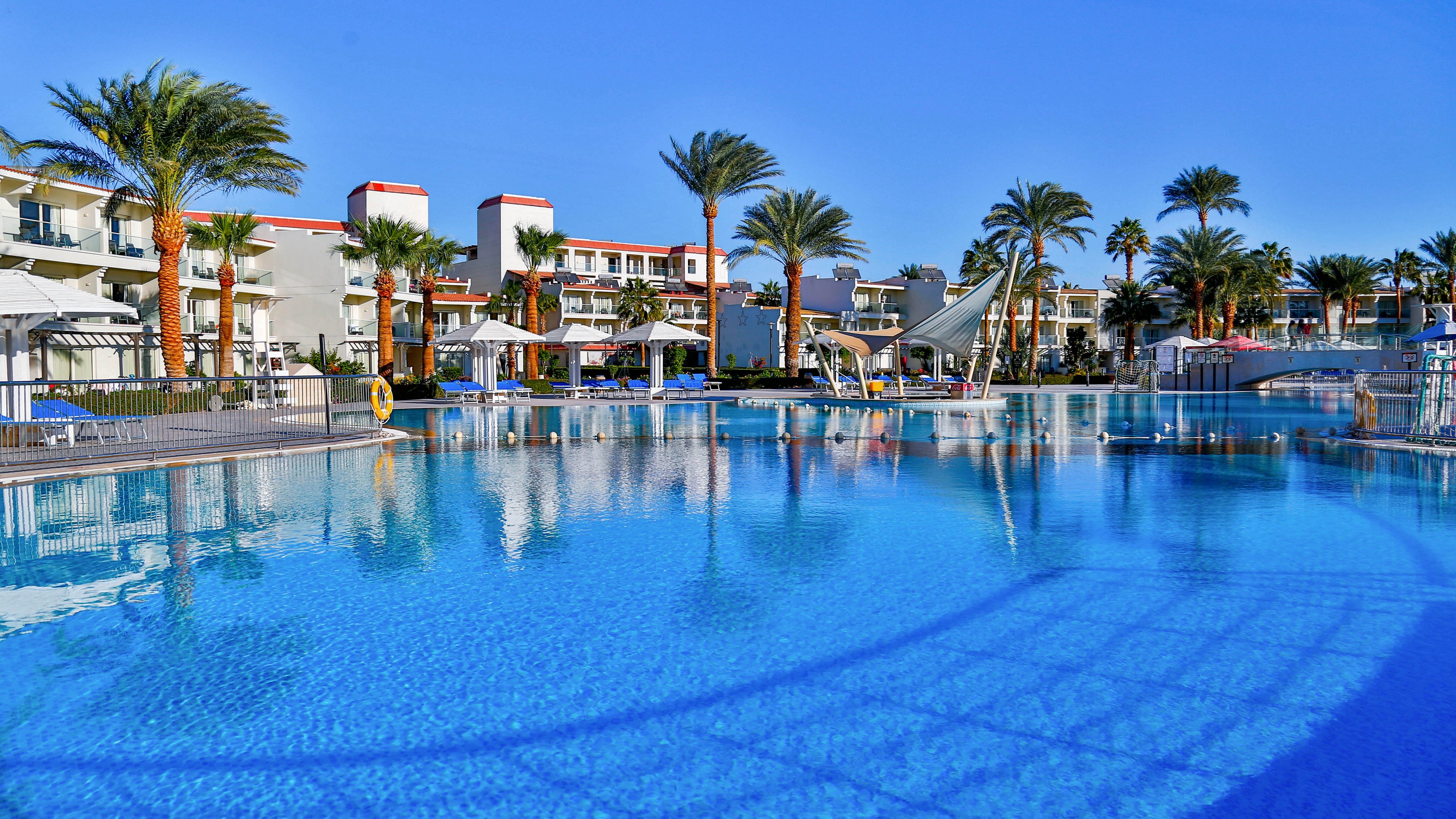 A large, clear blue swimming pool dominates the foreground of a sunny resort scene. Palm trees and multi-story white buildings with red accents line the poolside, suggesting a vacation hotel complex. Lounge areas, umbrellas, and pool safety equipment are visible around the water. The bright blue sky and reflections in the pool create a vibrant, relaxing atmosphere ideal for holidays.