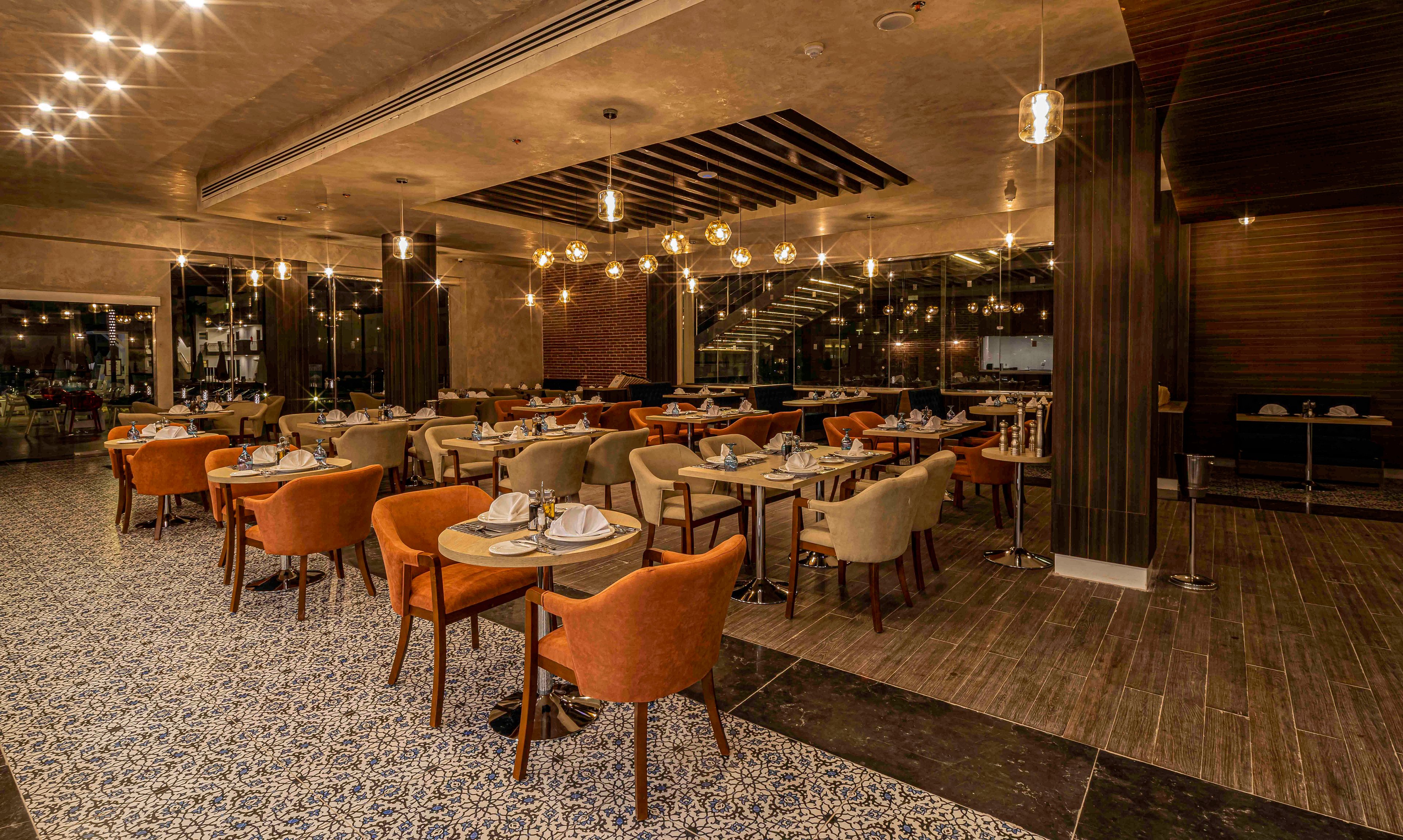 A contemporary restaurant dining area is shown with neatly arranged tables set for guests. Soft upholstered chairs in orange and beige surround wooden tables with plates and glassware. Warm pendant lights hang from the ceiling, reflecting in large windows and creating a cozy ambiance. The space features a mix of patterned tile flooring and dark wood, emphasizing a stylish, upscale atmosphere. No people are present, highlighting the interior design and layout.