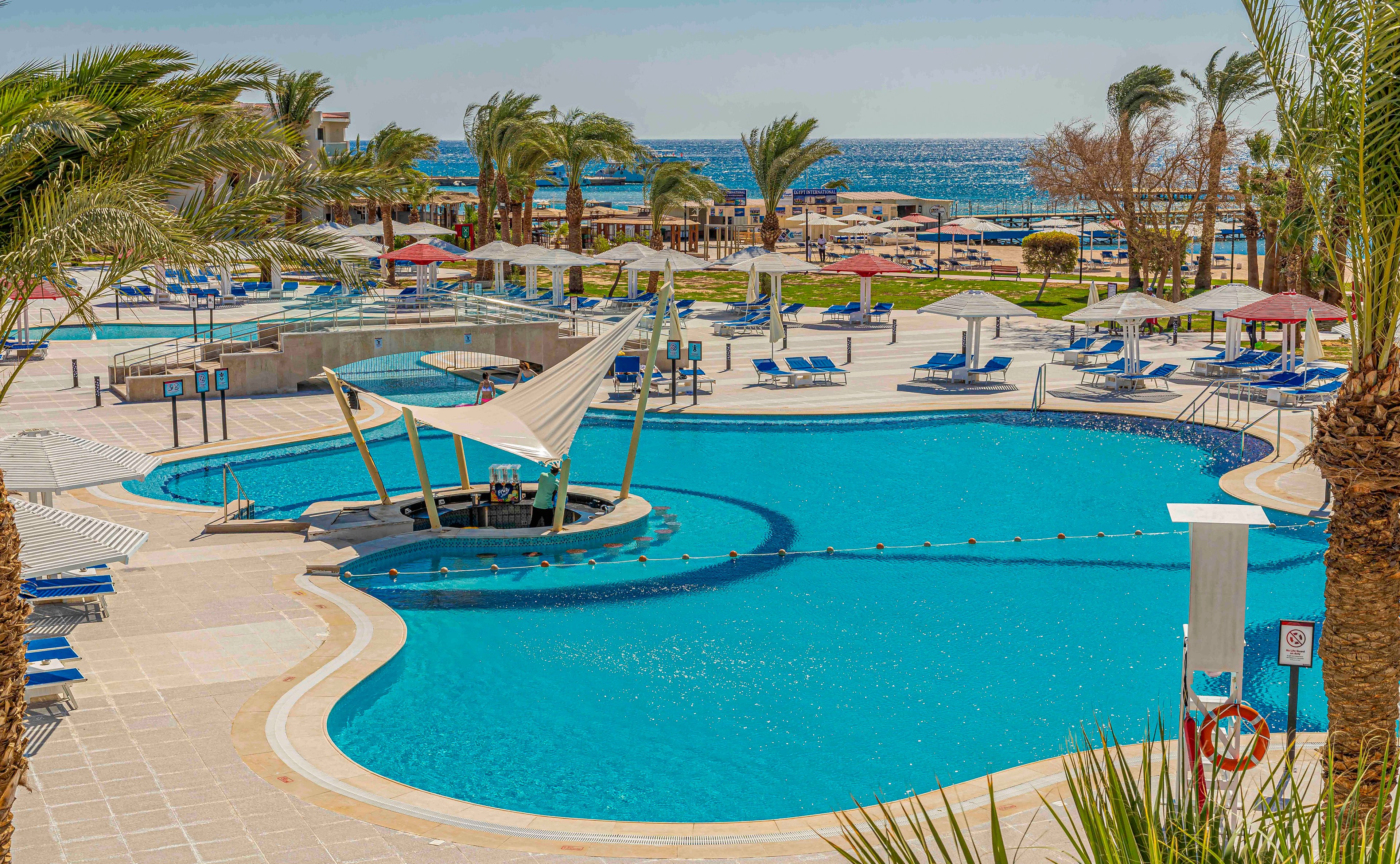 Large curved swimming pools dominate this sunny seaside resort scene. Blue loungers and umbrellas surround the pool deck, with palm trees adding a tropical atmosphere. A shaded seating structure stands in the water, and safety equipment is visible near the pool edge. In the background, the sea and additional sunbeds extend toward the horizon under a clear sky.