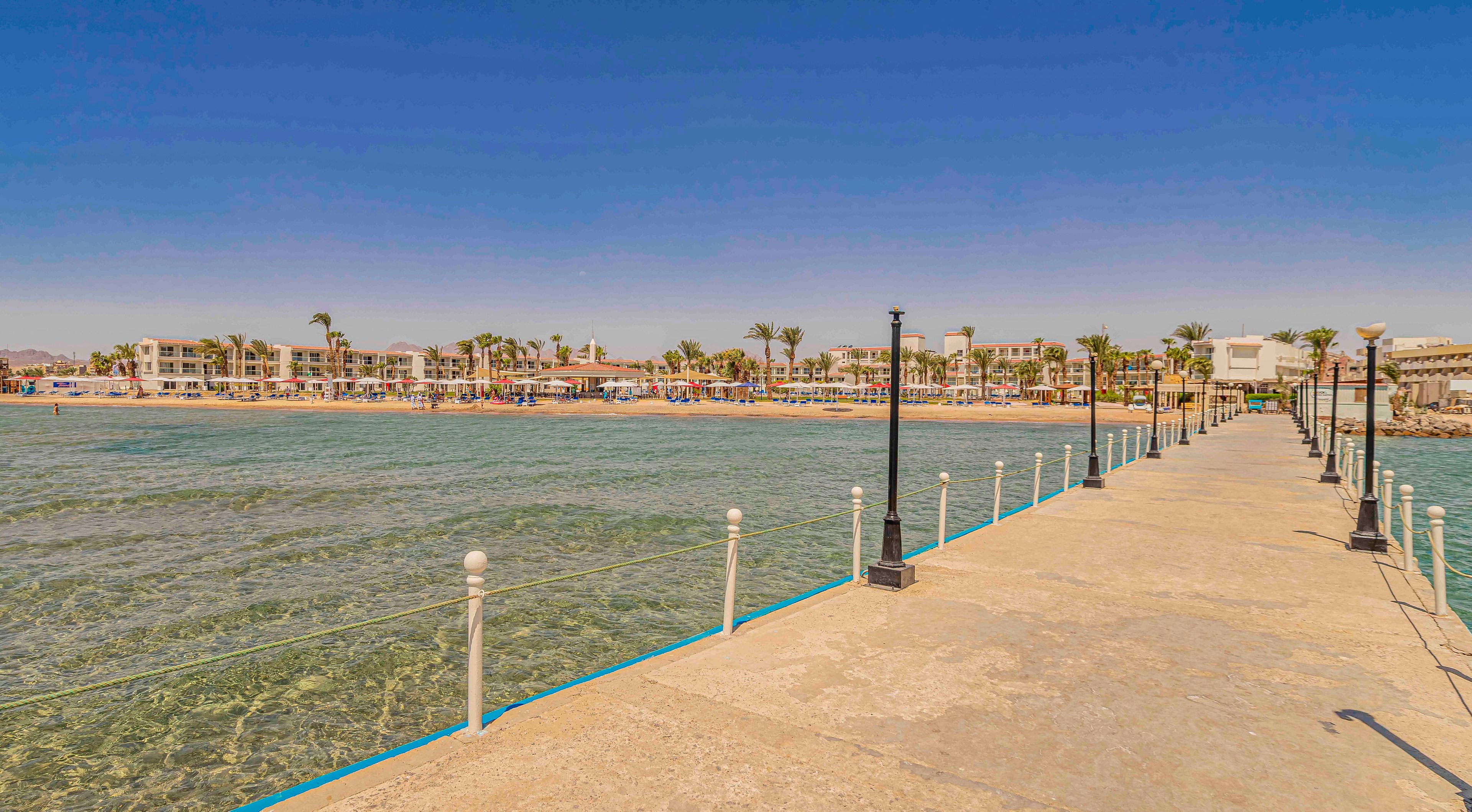 A long concrete pier extends over shallow, clear water toward a distant seaside resort. Low-rise buildings and palm trees line the shoreline under a bright blue sky. The calm sea reveals underwater textures near the surface. White posts and ropes run along the pier, guiding the eye toward the beach. The overall scene suggests a warm, tranquil vacation destination.
