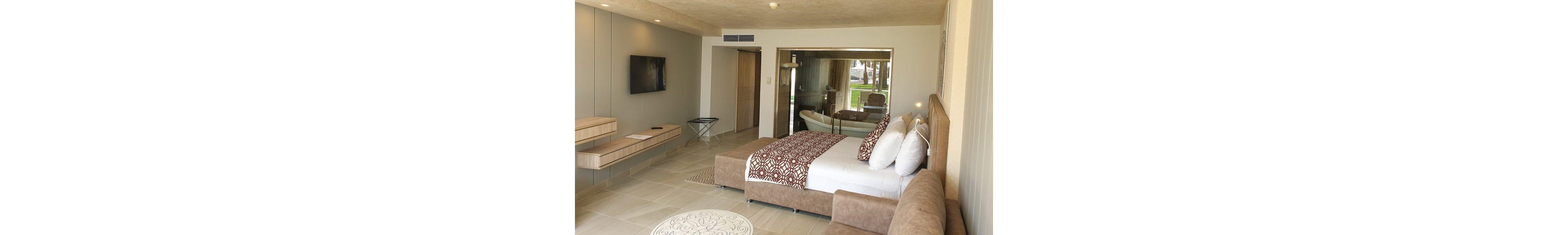 Contemporary hotel room featuring a large bed with patterned brown and white linens and multiple pillows. A glass wall reveals a freestanding bathtub and bathroom area with views to the outdoors. The room includes a mounted flat-screen TV, floating wooden shelves, and a small luggage rack. Neutral beige and brown tones, tiled flooring, and minimal decor create a clean, modern atmosphere.