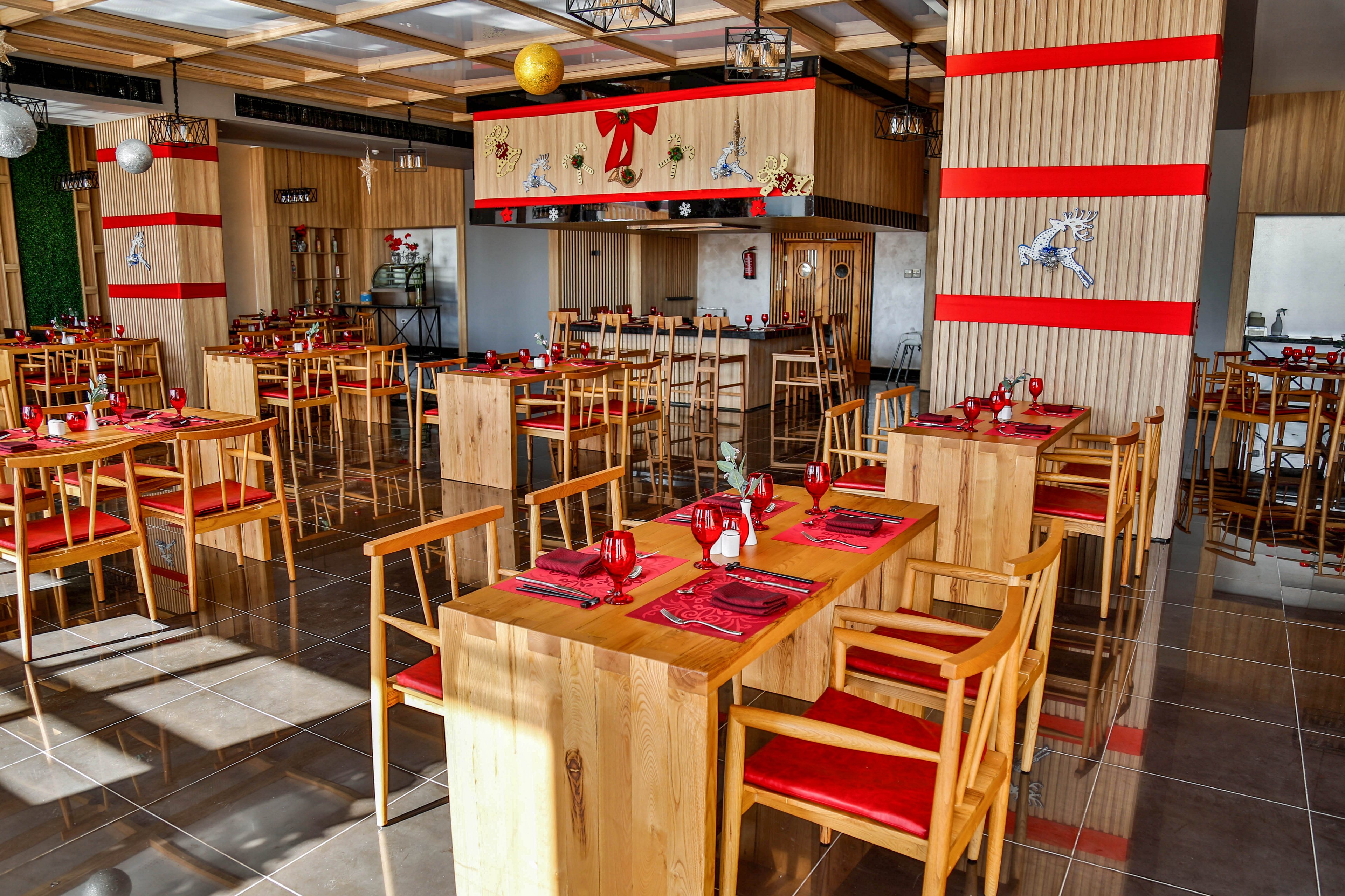 A modern restaurant dining area features light wooden tables and chairs with red seat cushions. Each table is neatly set with red placemats, folded napkins, and glassware, suggesting readiness for guests. The space is decorated with red ribbons, bows, and hanging ornaments, creating a festive atmosphere. Large windows and reflective flooring enhance the bright, spacious feel of the interior.