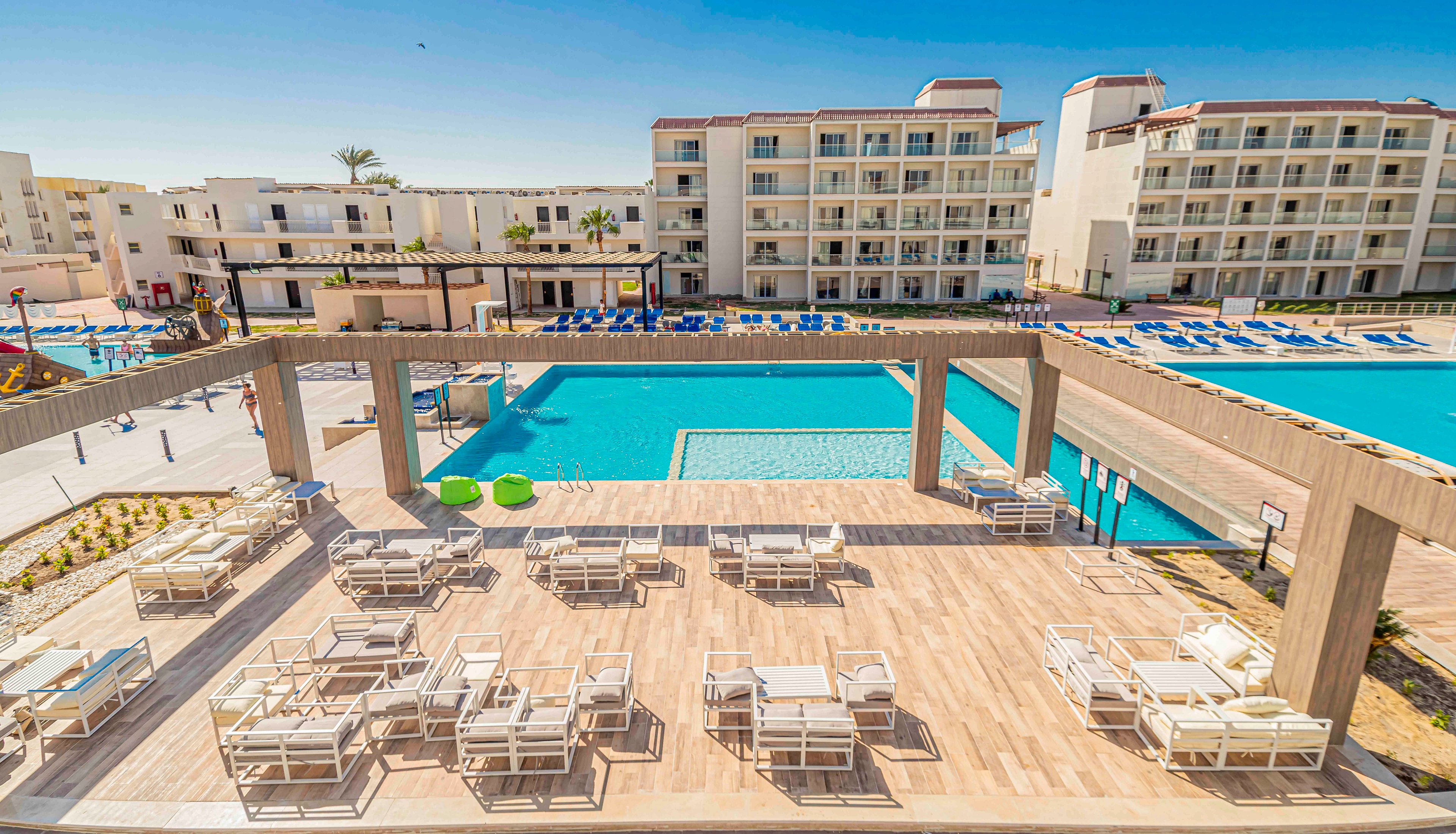A spacious resort terrace overlooks a large outdoor swimming pool surrounded by modern hotel buildings. White lounge chairs and tables are neatly arranged on the wooden deck under open pergola structures. Blue sunbeds line the far side of the pool, adding a vibrant contrast to the neutral architecture. The scene is brightly lit under a clear blue sky, suggesting a warm, sunny vacation destination.