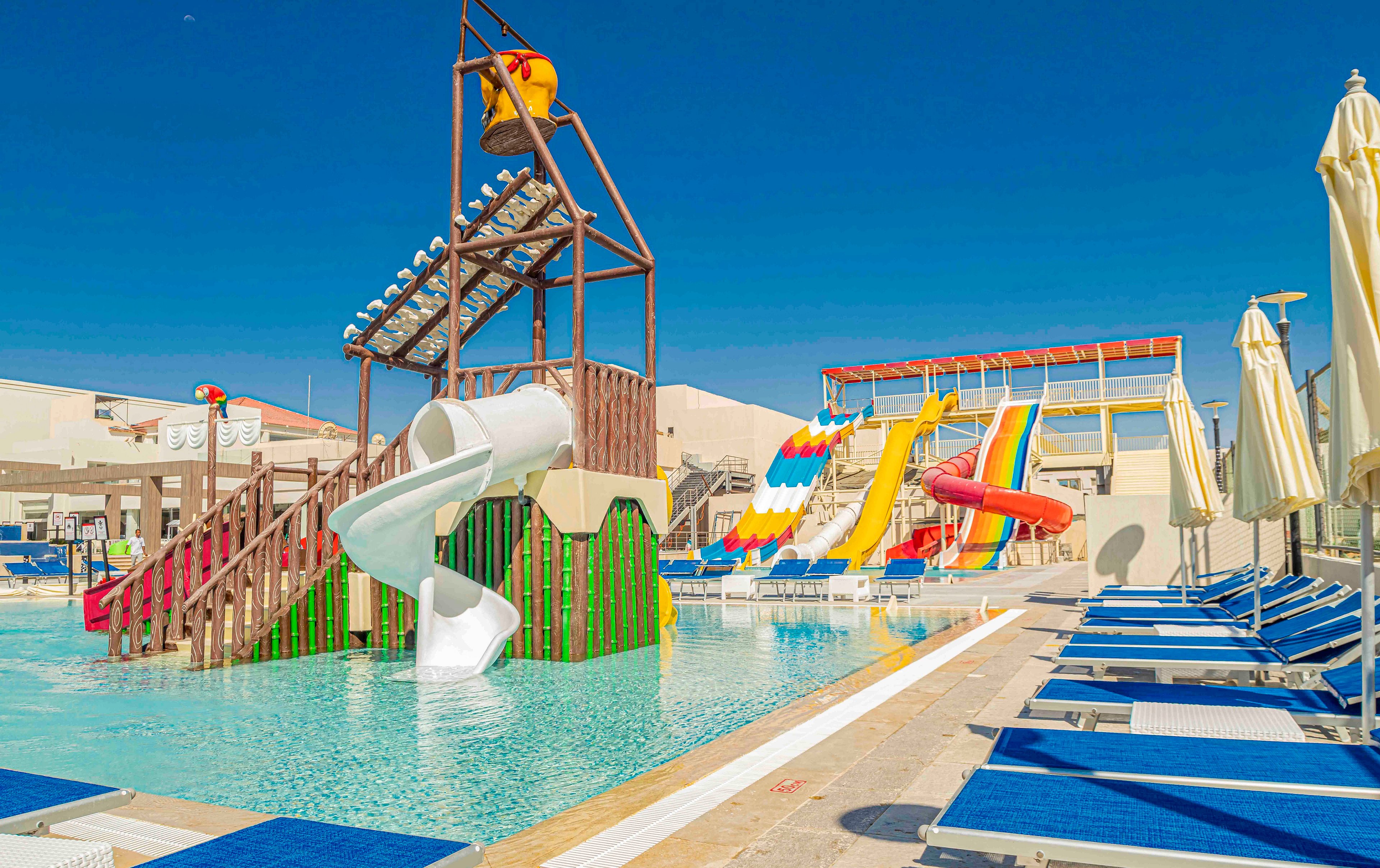 An outdoor resort water park features multiple colorful slides empty under a clear blue sky. A white spiral slide and wooden play structure rise from a shallow pool with calm turquoise water. In the background, tall rainbow slides and a tipping water bucket add to the fun atmosphere. Blue sun loungers and closed umbrellas line the poolside deck, suggesting a relaxing vacation setting.