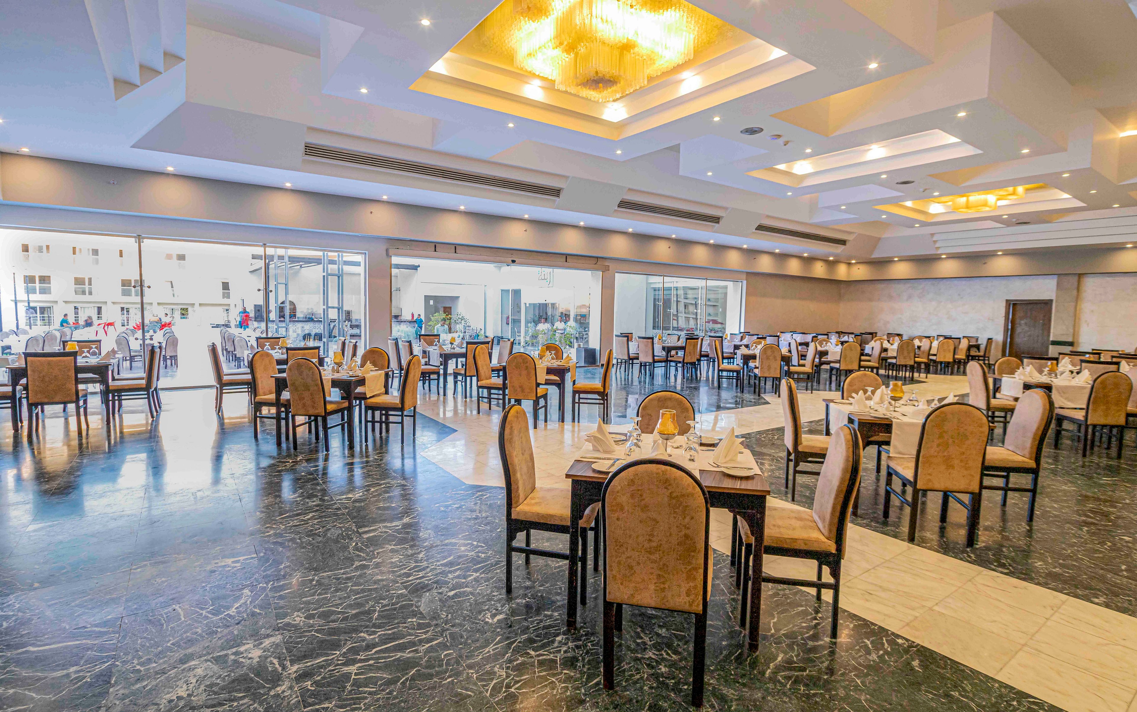 A large, modern restaurant dining hall features neatly arranged tables and upholstered chairs. The space has a polished marble-style floor and a bright ceiling with recessed lighting and chandeliers. Place settings with plates, glasses, and folded napkins are prepared on several tables, suggesting readiness for guests. Large windows and glass doors along one side let in natural light and offer views of the outdoor area.