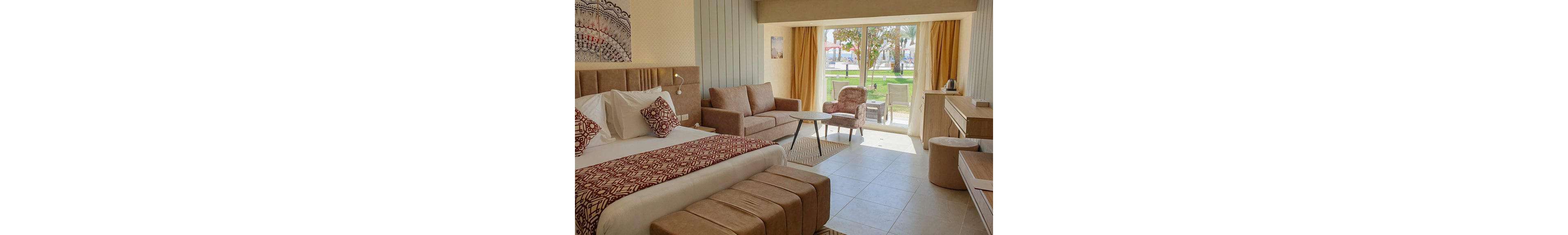 A spacious hotel room features a large bed with patterned red and white textiles and matching cushions. A comfortable seating area with a sofa, armchair, and small table faces a wide glass door. The door opens to a terrace with outdoor chairs and a view of greenery and palm trees. Neutral beige and tan tones dominate the interior, creating a warm, relaxed atmosphere. Natural light floods the room, highlighting the modern, tidy decor.