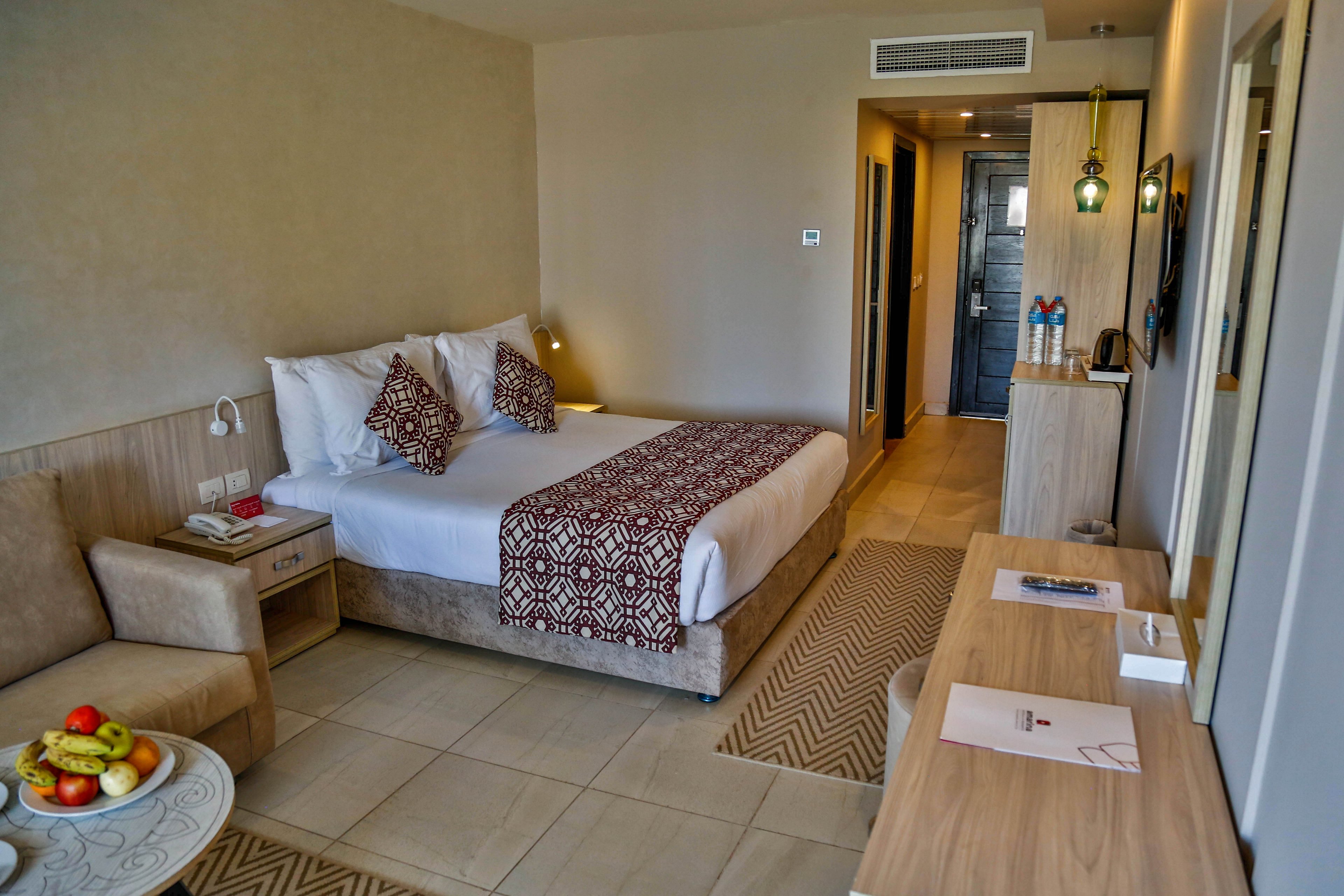 A contemporary hotel room features a neatly made double bed with patterned pillows and runner. The space includes a light wood desk, a small sofa, and a side table with a bowl of fresh fruit. Warm lighting and neutral tones create a calm, inviting atmosphere. A hallway leads to the entrance door and bathroom area, emphasizing the room’s spacious layout and modern amenities.