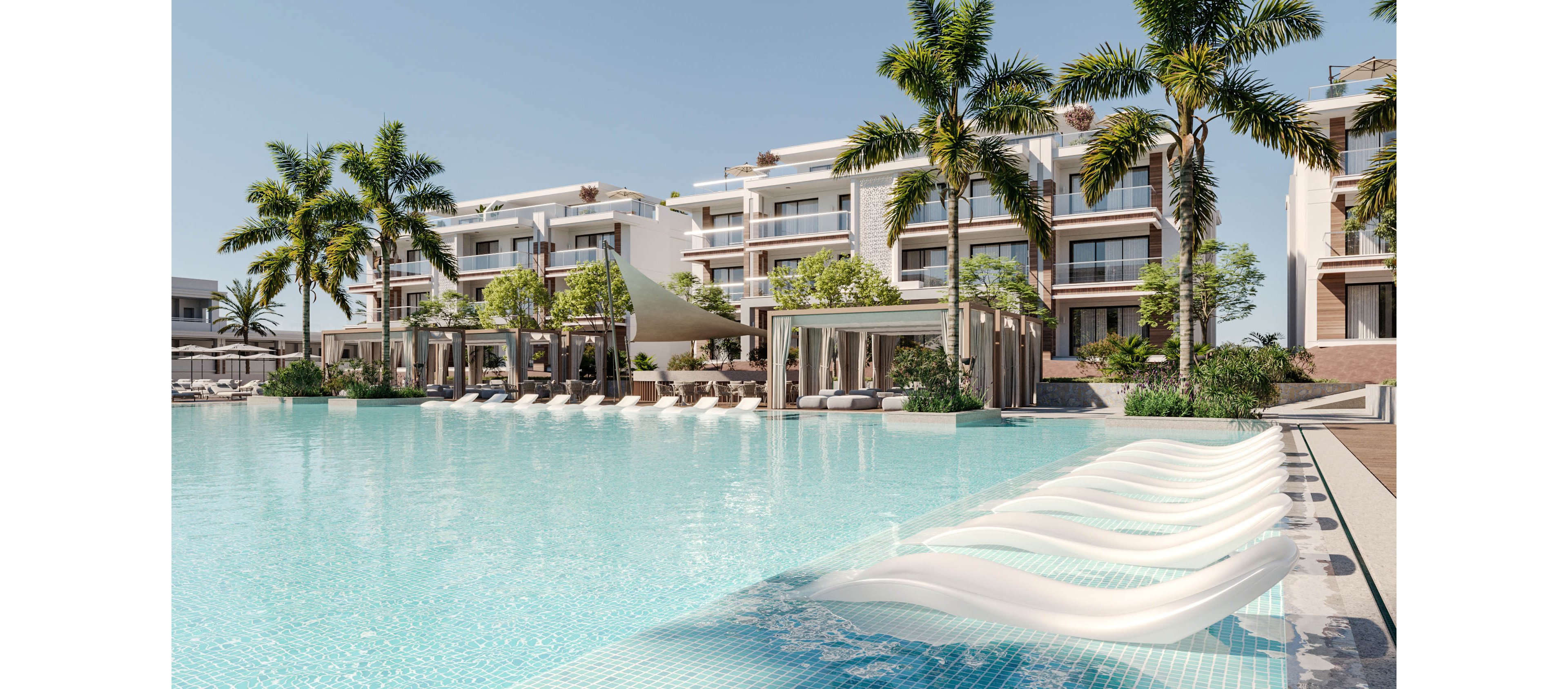 A modern luxury resort features a large, inviting swimming pool in the foreground. White in-water loungers line the shallow edge, facing contemporary multi-story hotel buildings. Palm trees and lush greenery surround shaded cabanas and seating areas, creating a tropical atmosphere. The bright daylight and clear sky emphasize a calm, relaxing vacation setting.
