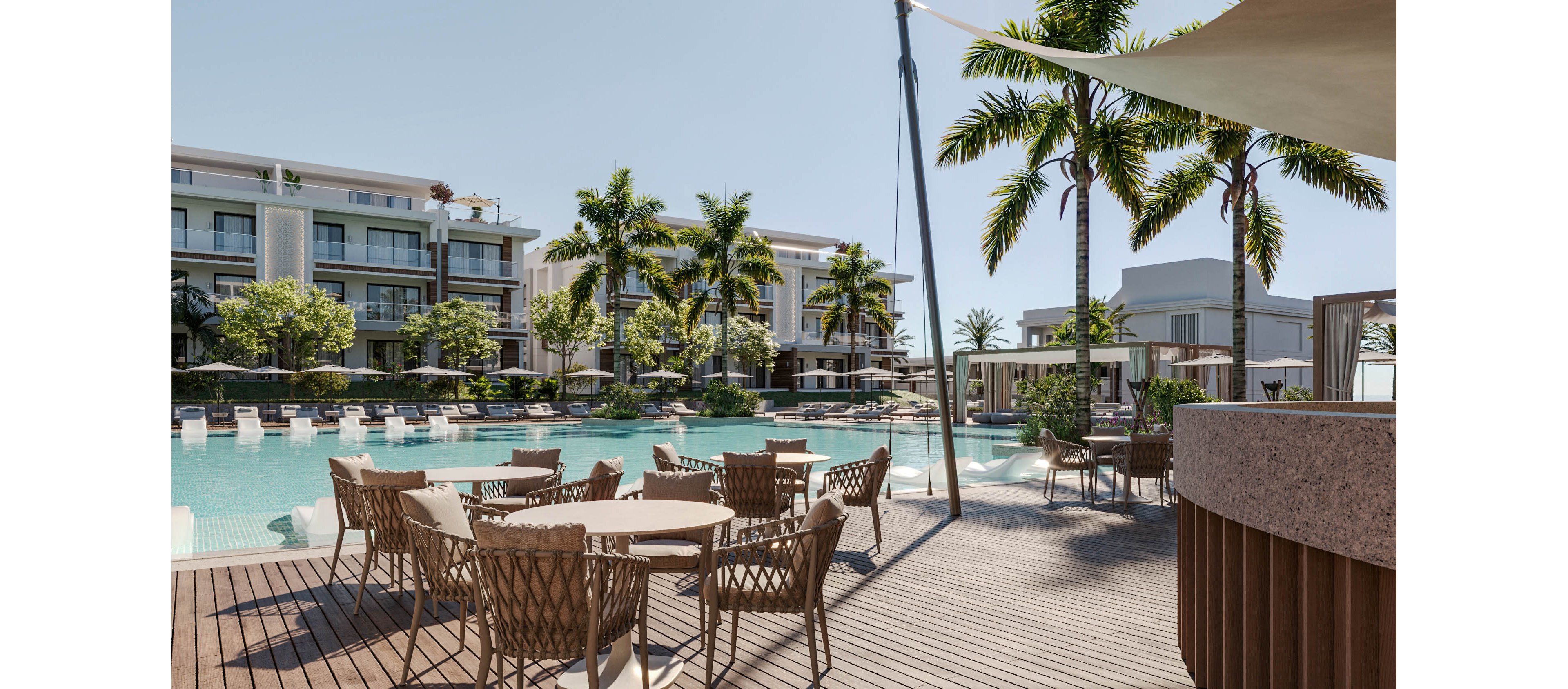 An upscale resort pool area features woven chairs and round tables arranged on a wooden deck. Sun loungers line the edge of a large swimming pool, facing modern multi-story buildings in the background. Tall palm trees and lush greenery create a tropical atmosphere under clear daylight. The scene appears calm and inviting, suggesting a relaxing vacation or leisure setting.