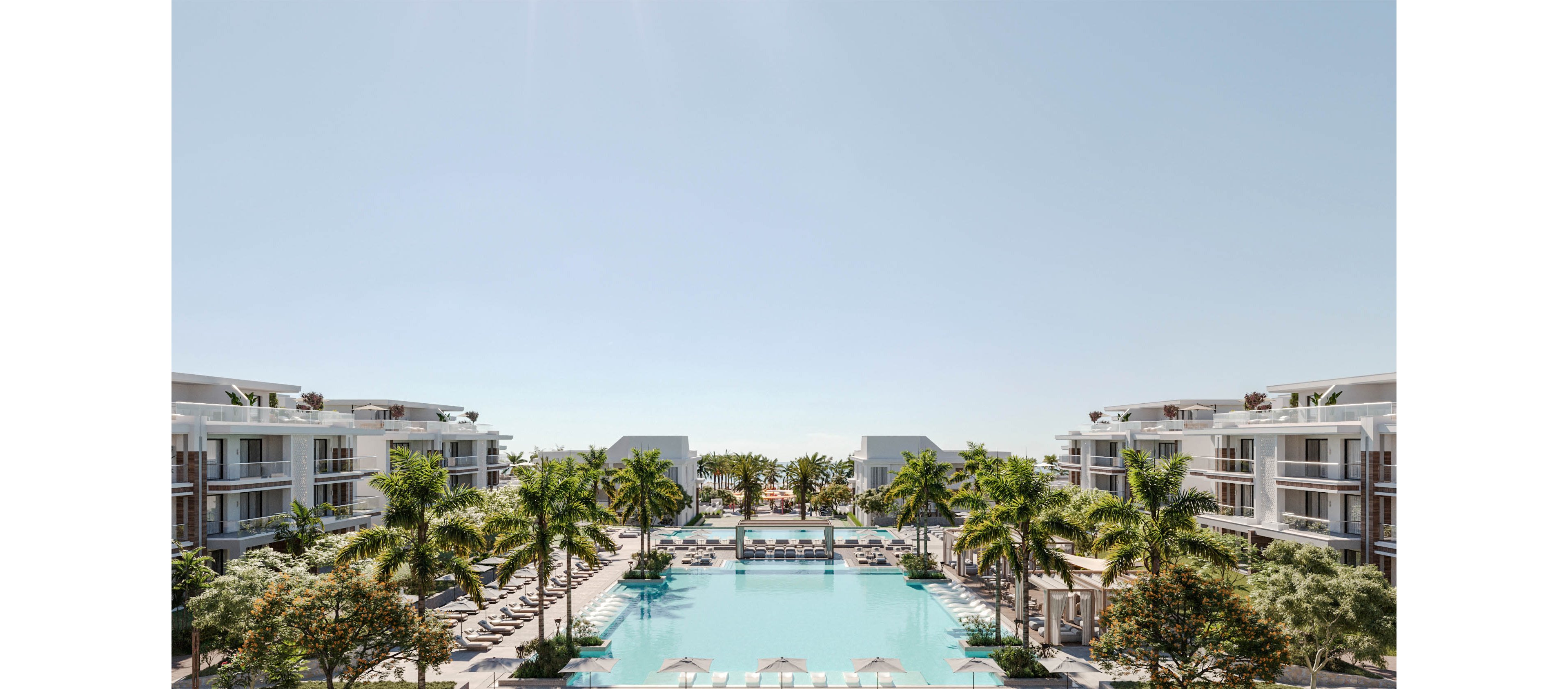 A contemporary resort complex features a large central swimming pool framed by palm trees. White multi-story buildings with balconies line both sides, creating a symmetrical composition. Lounge chairs and umbrellas surround the pool, suggesting a relaxing vacation atmosphere. The bright daylight and clear sky emphasize a warm, tropical climate and luxurious outdoor setting.