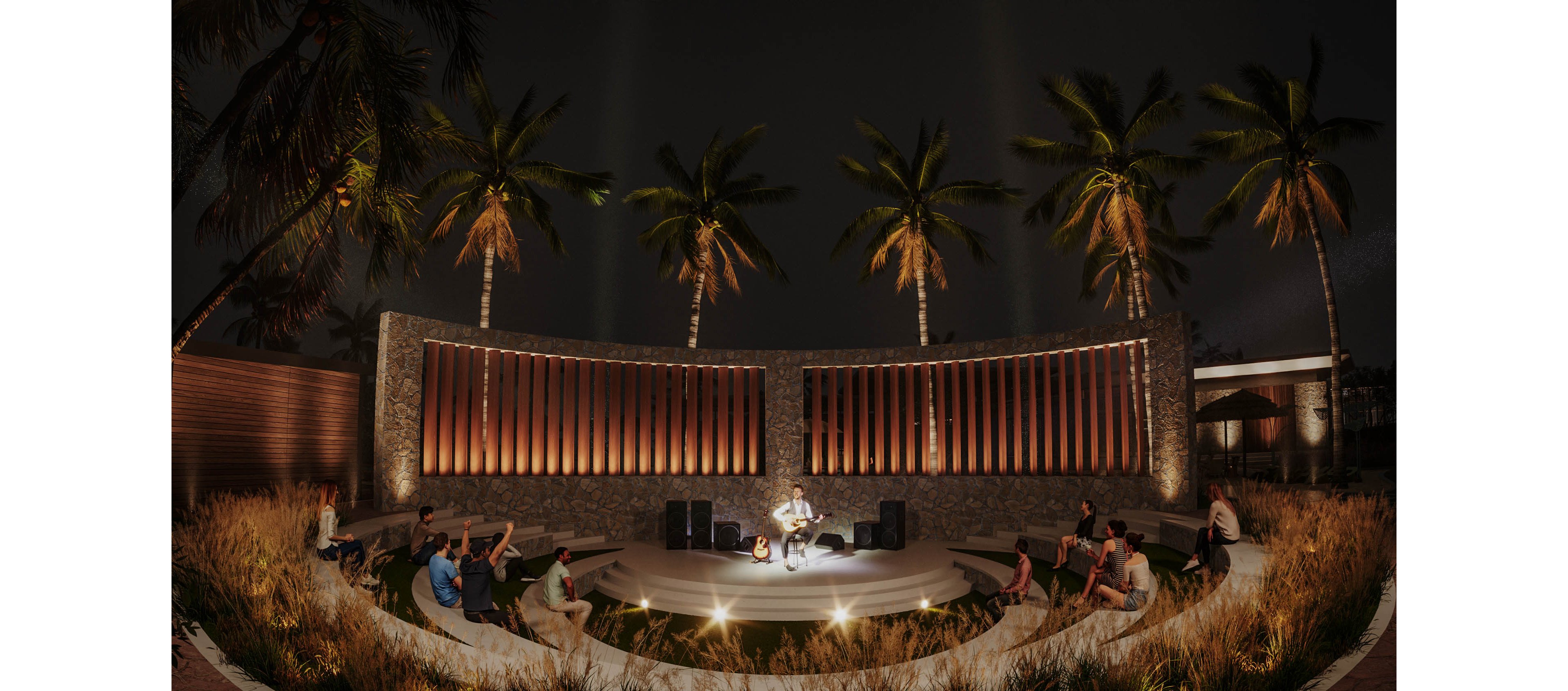 A musician performs on a circular stage in an intimate outdoor amphitheater at night. Audience members sit on curved stone benches surrounded by ornamental grasses and soft ground lighting. Tall palm trees rise behind a modern textured wall, creating a tropical resort atmosphere. Warm spotlights highlight the performer and stage equipment, contrasting with the dark night sky.