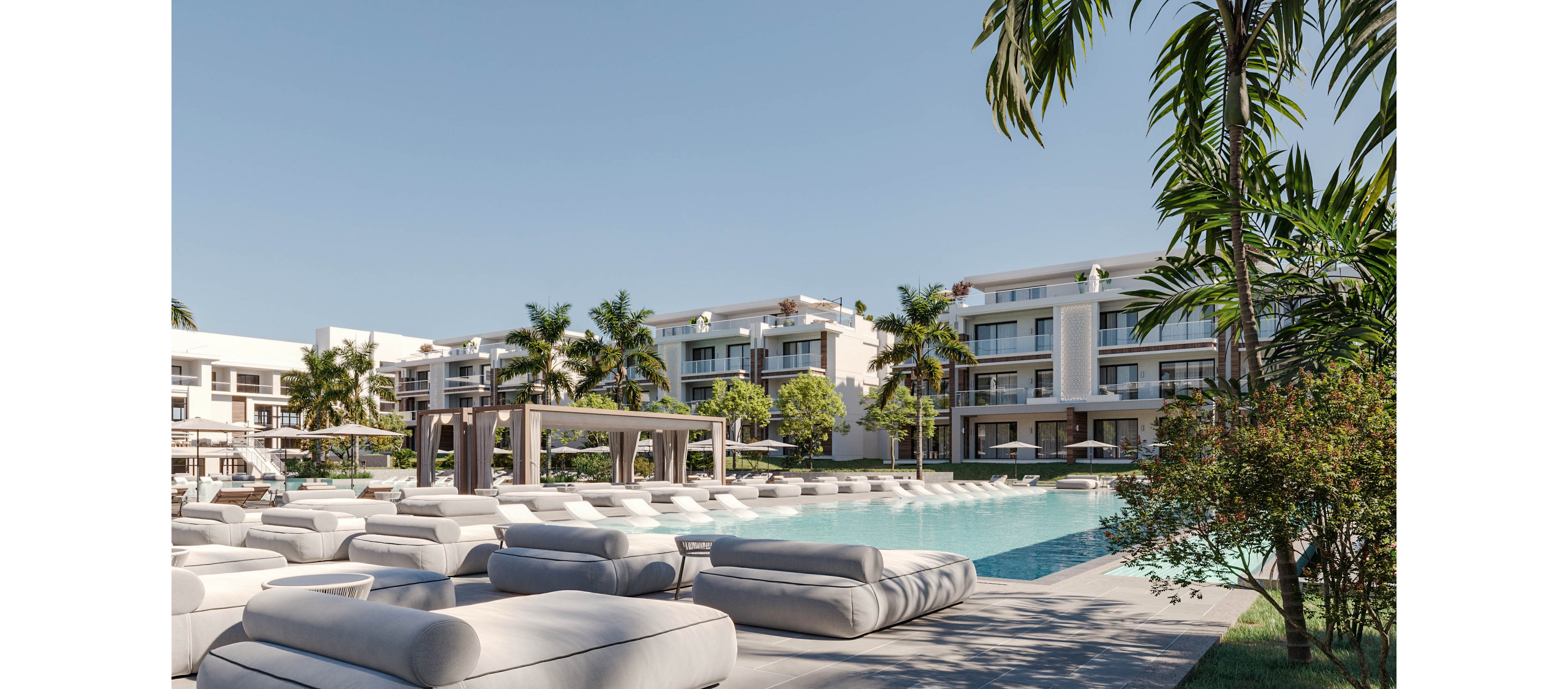 A contemporary resort features a large outdoor swimming pool surrounded by plush white sun loungers. Low-rise modern buildings with balconies frame the pool area, creating an upscale vacation setting. Tall palm trees and manicured greenery add a tropical atmosphere under a clear blue sky. The scene suggests relaxation, leisure, and resort-style living in a sunny destination.