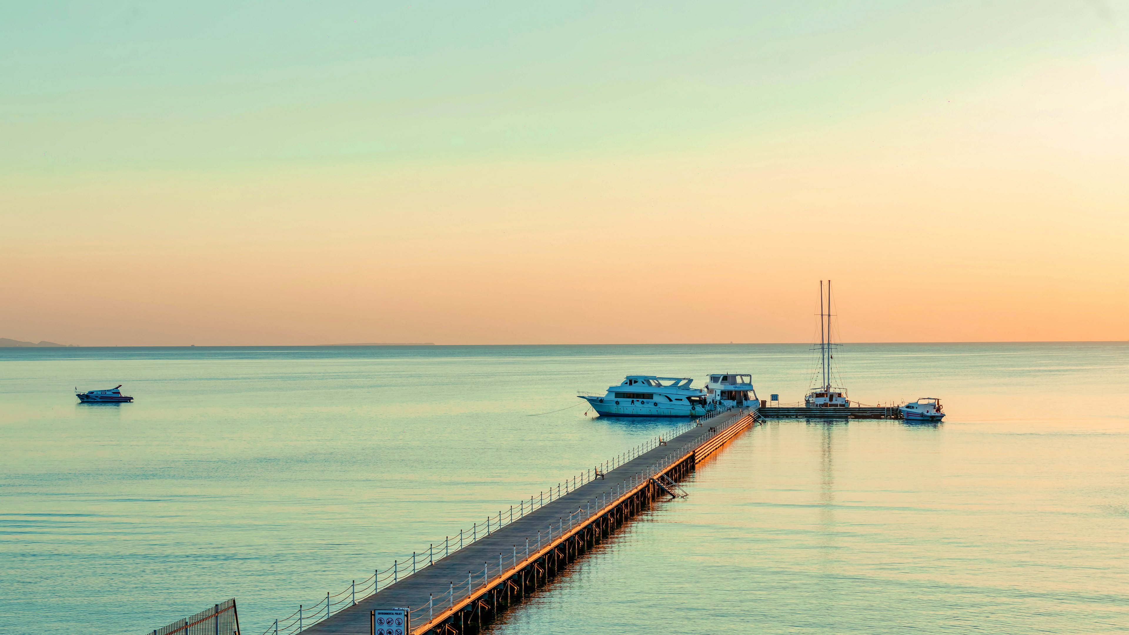 A narrow wooden pier extends into calm, open water toward several moored boats. The scene is set during sunset or sunrise, with soft pastel colors reflecting on the sea. The horizon is clear and distant, creating a peaceful, minimalist coastal landscape. Gentle light and smooth water emphasize a serene, relaxing atmosphere.