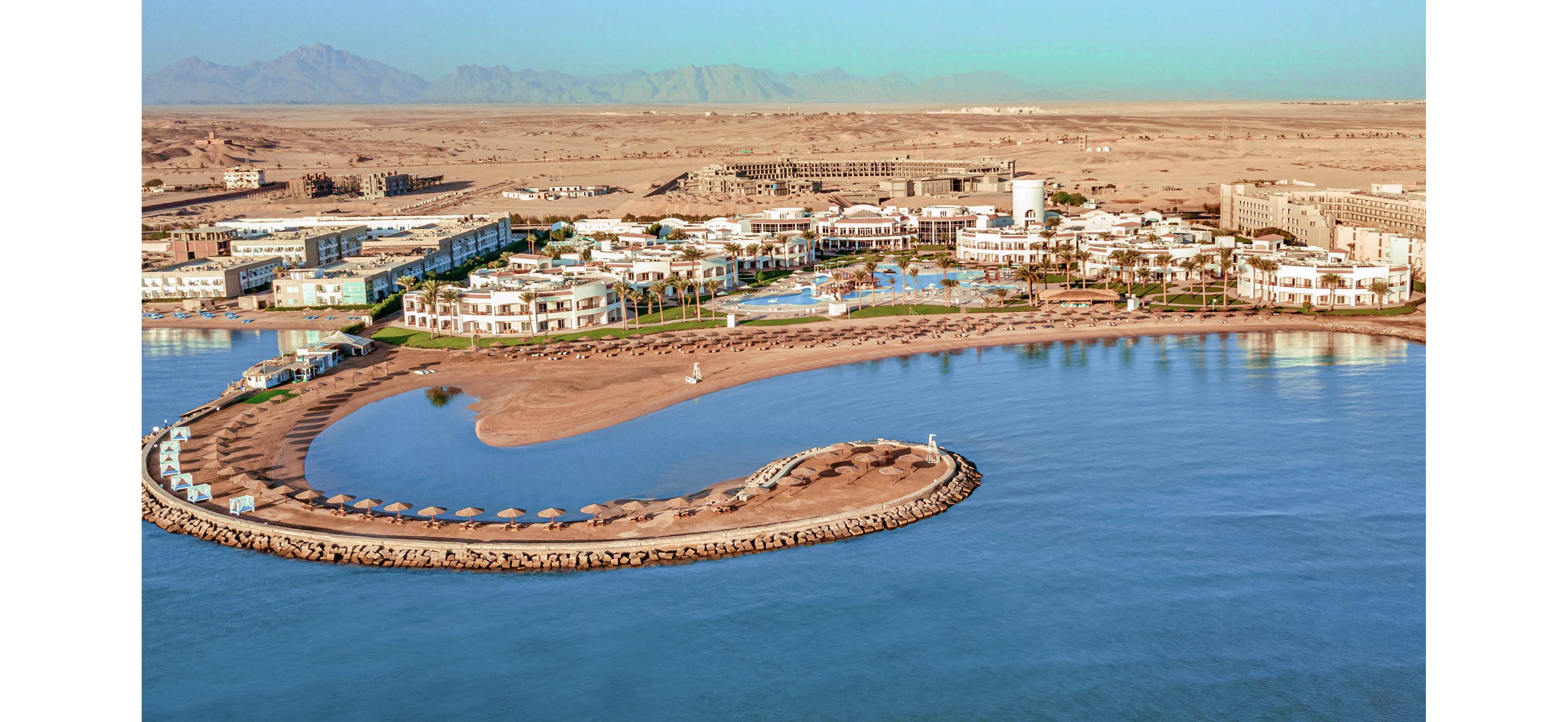 A luxurious seaside resort stretches along a curved artificial peninsula surrounded by calm turquoise water. The aerial perspective reveals hotel buildings, pools, palm-lined walkways, and a sandy beach with evenly spaced umbrellas. Beyond the resort, a vast desert landscape extends to distant mountain ranges under a clear blue sky. The scene conveys a tranquil vacation destination contrasting vivid blue sea with warm sandy tones.