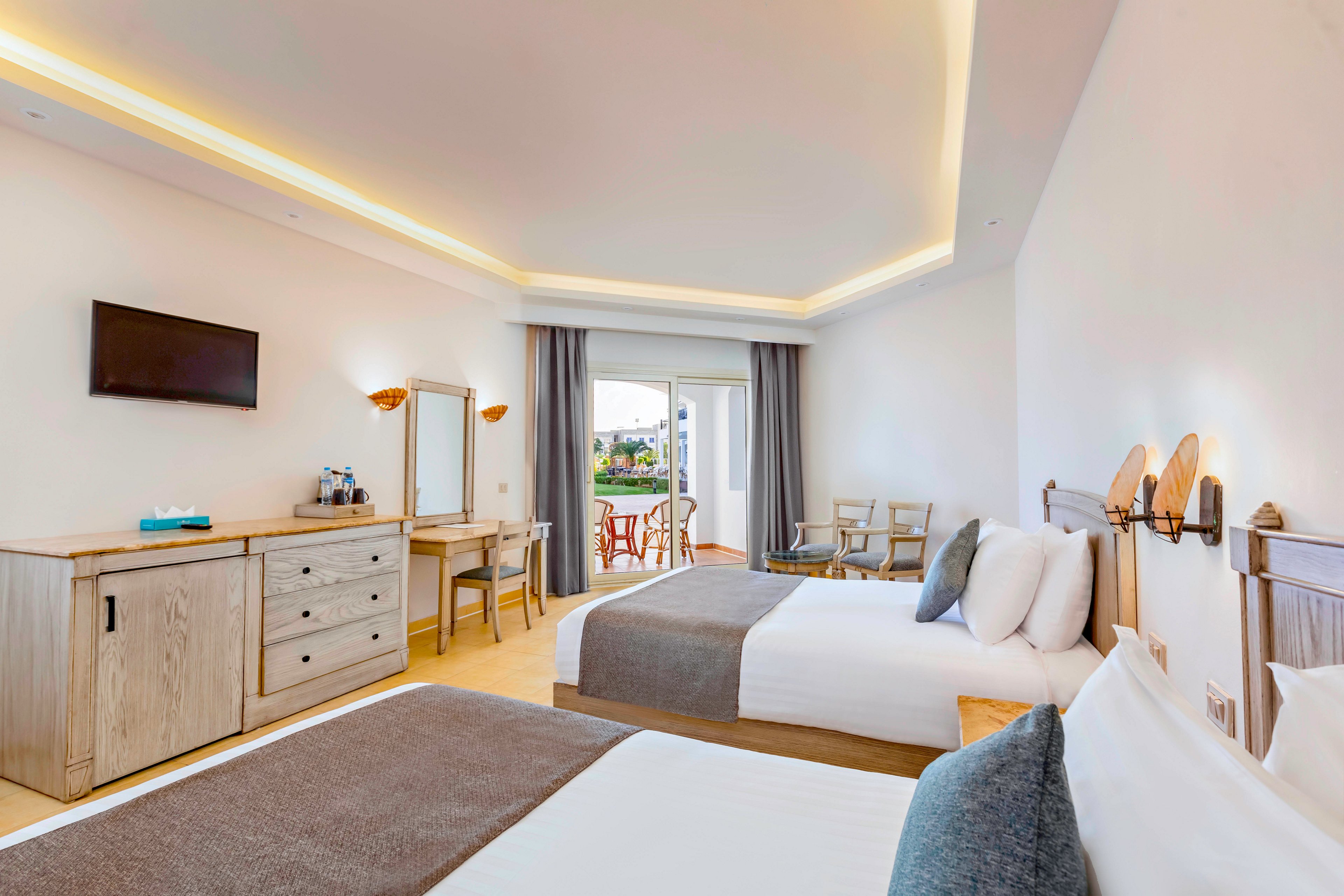 A contemporary hotel room features two neatly made double beds with neutral bedding and accent pillows. Light wood furniture, including a dresser, desk, and mirror, lines the wall beneath a mounted flat-screen TV. Warm recessed ceiling lighting and wall sconces create a welcoming atmosphere. At the far end, large glass doors open to a balcony with outdoor chairs and a view of greenery.