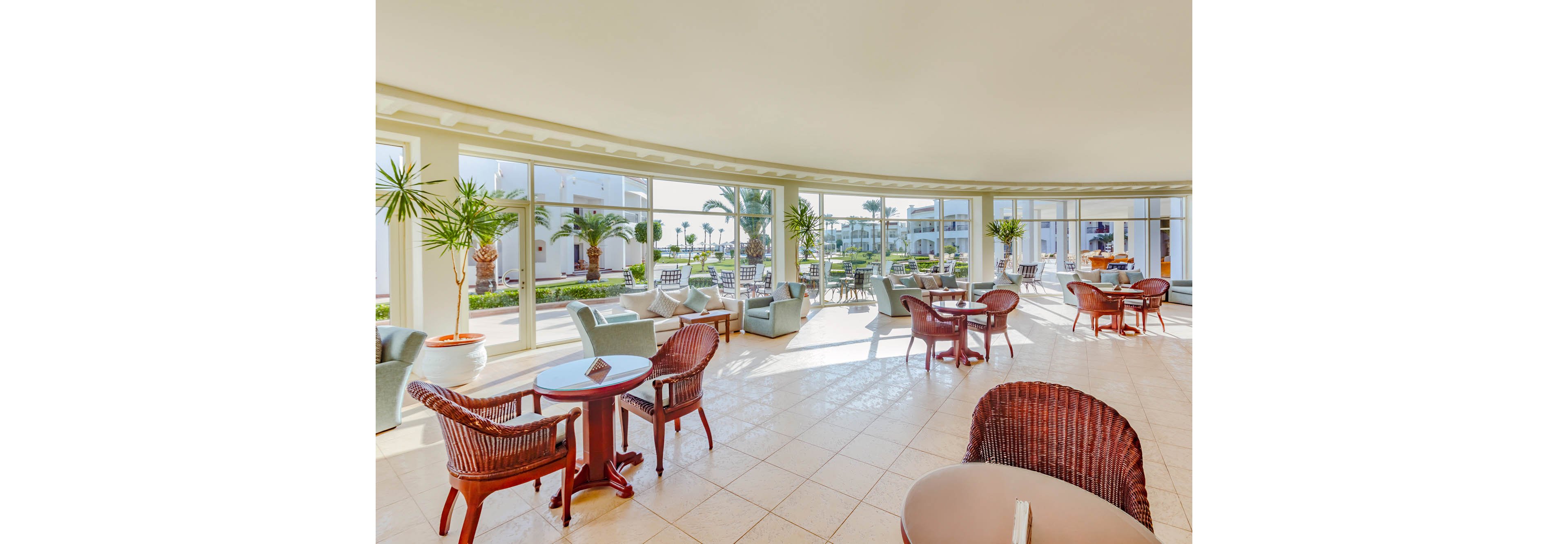 Spacious hotel lounge area features wicker chairs and round wooden tables arranged on a tiled floor. Large floor-to-ceiling windows open onto a sunny garden with palm trees and white resort buildings. Soft natural light fills the room, creating an airy and relaxed atmosphere ideal for casual meetings or quiet relaxation. The interior decor is contemporary with neutral tones and touches of greenery in potted plants.