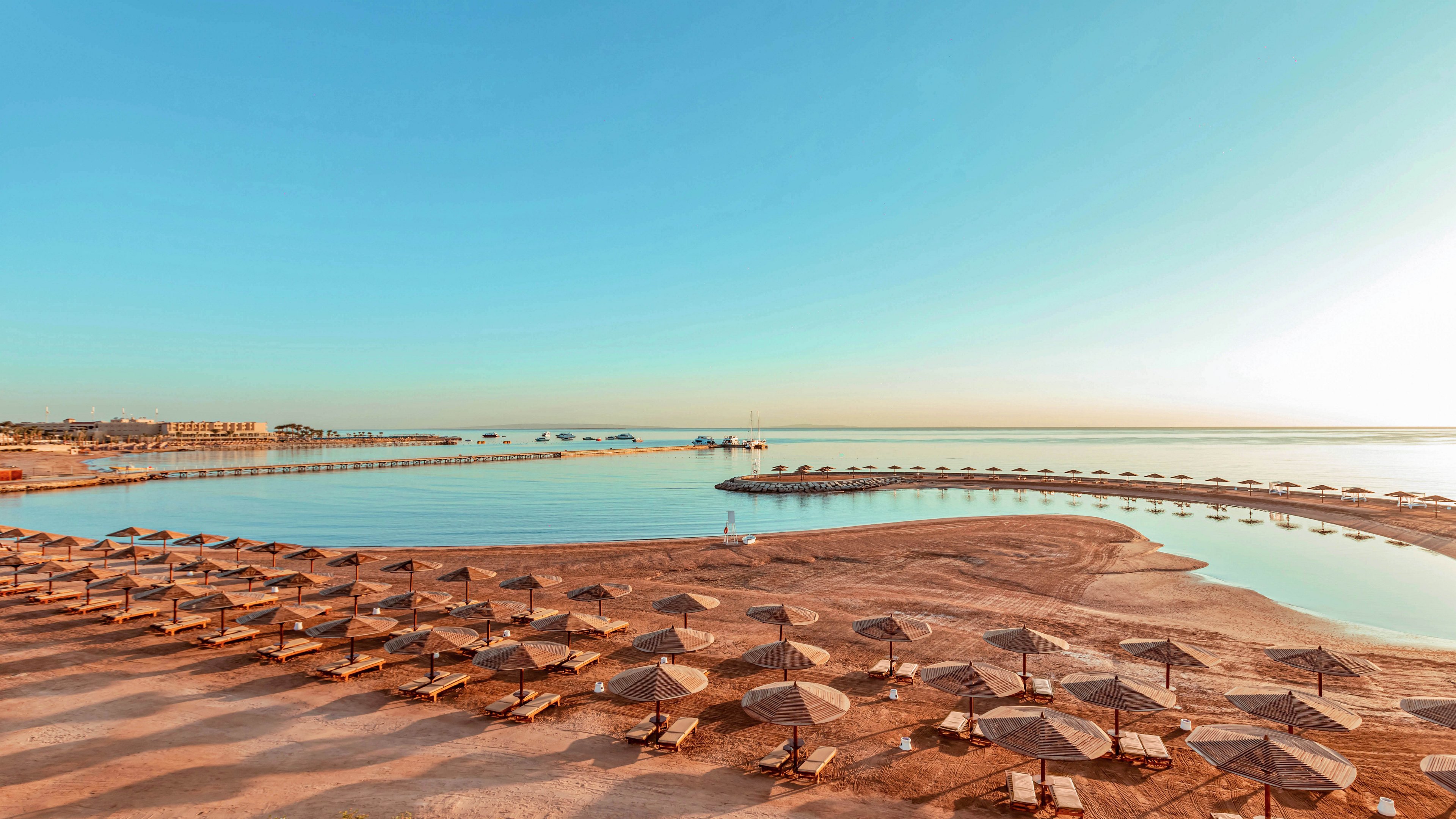 A wide sandy beach is lined with rows of sun umbrellas and loungers facing a calm, clear sea. Curved piers extend into the water, creating sheltered swimming areas and adding structure to the coastline. The sky is bright and cloudless, casting warm golden light across the sand and water. In the distance, low resort buildings and additional beach facilities are visible along the shore.