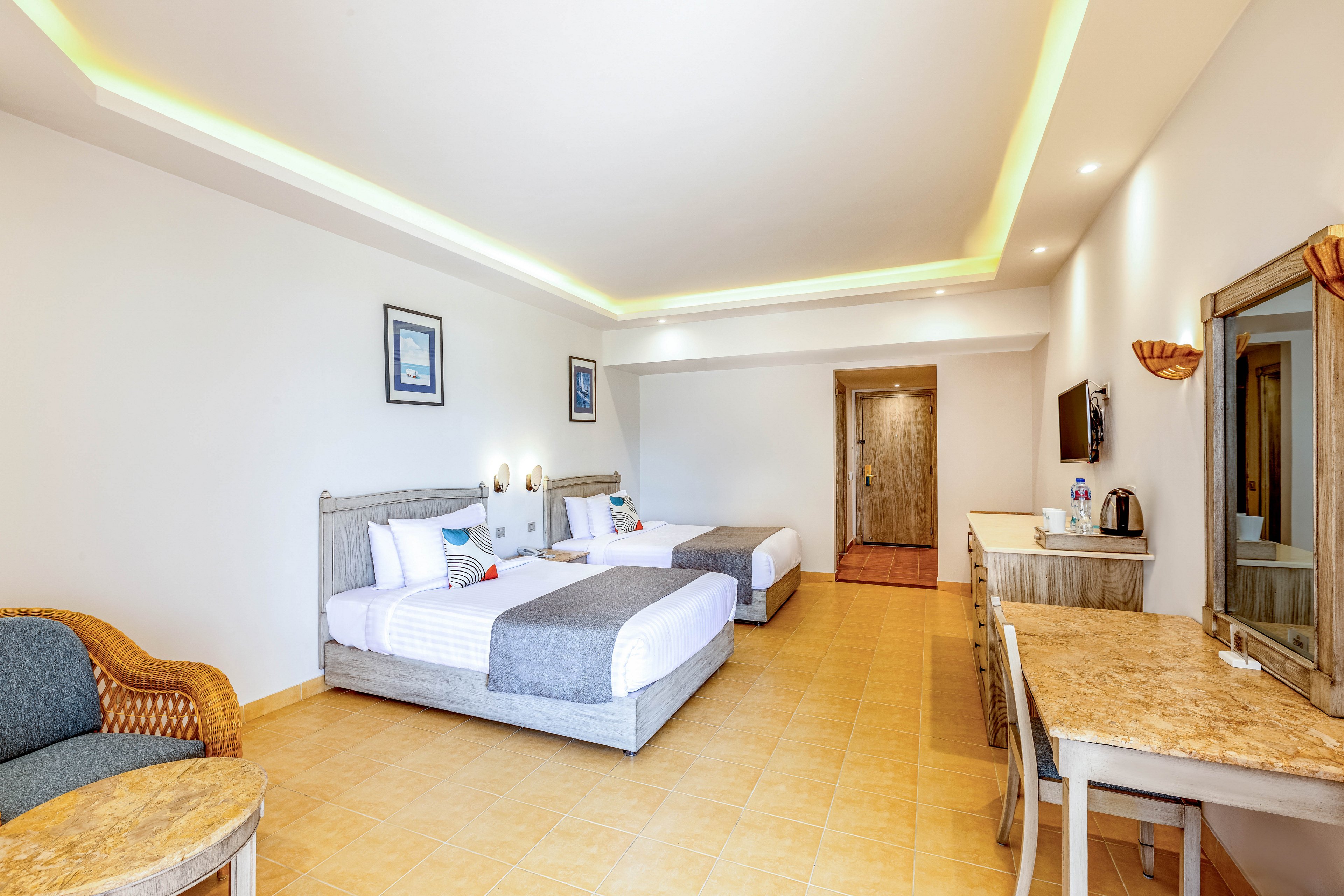 Spacious hotel guest room featuring two neatly made twin beds with white linens and gray runners. The space includes a wicker armchair, a long desk with mirror, and a wall-mounted television. Warm tile flooring and recessed ceiling lighting create a bright, inviting atmosphere. Decorative pillows and framed artwork add subtle color accents to the clean, modern interior.