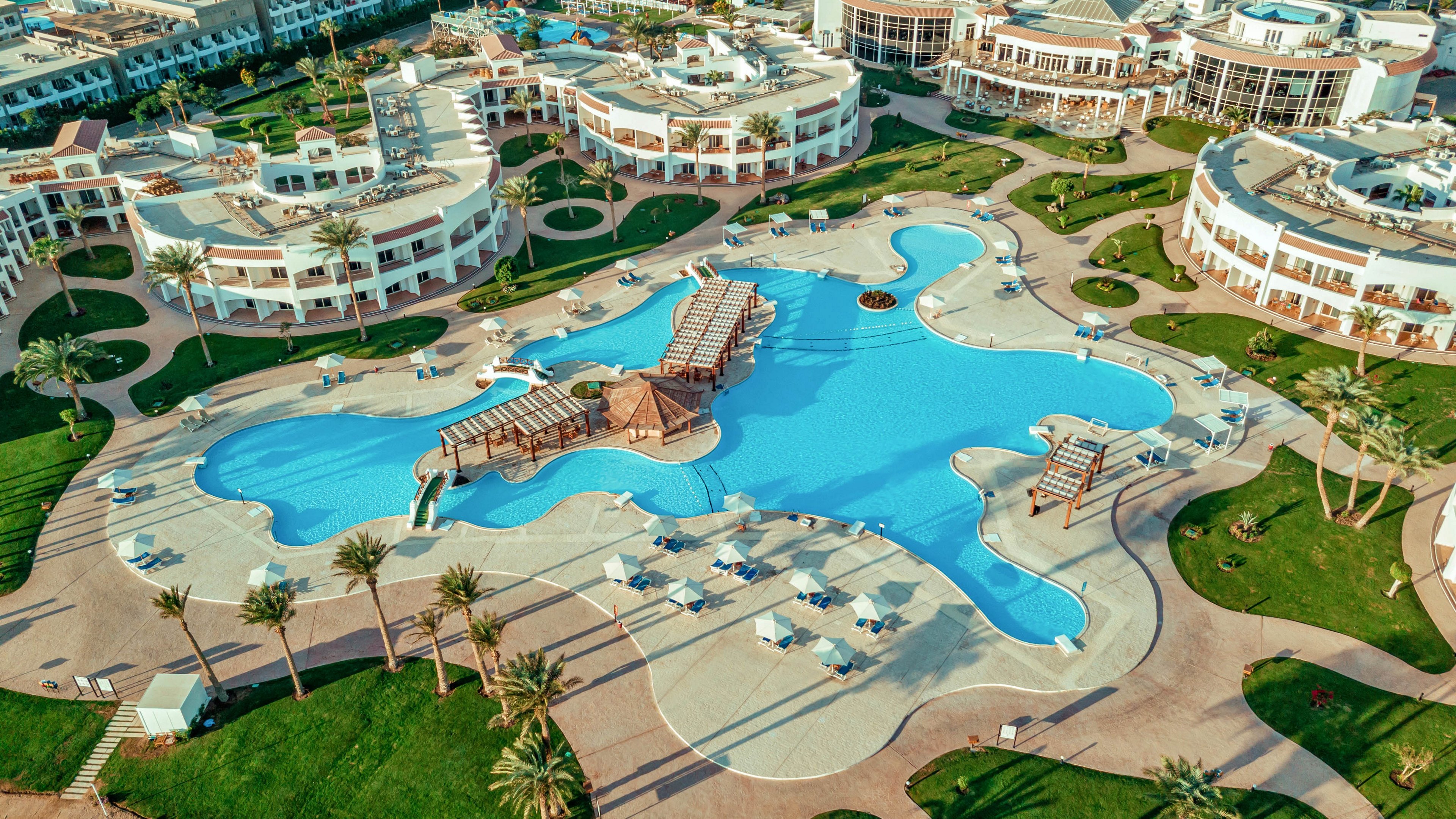 A large, irregularly shaped blue swimming pool sits at the center of a luxury resort complex. Curved white buildings with balconies surround the pool, creating a symmetrical layout. Sun loungers and umbrellas are arranged neatly around the poolside terraces, with wooden bridges crossing over the water. Palm trees and landscaped paths add a tropical atmosphere to the sunny outdoor setting.