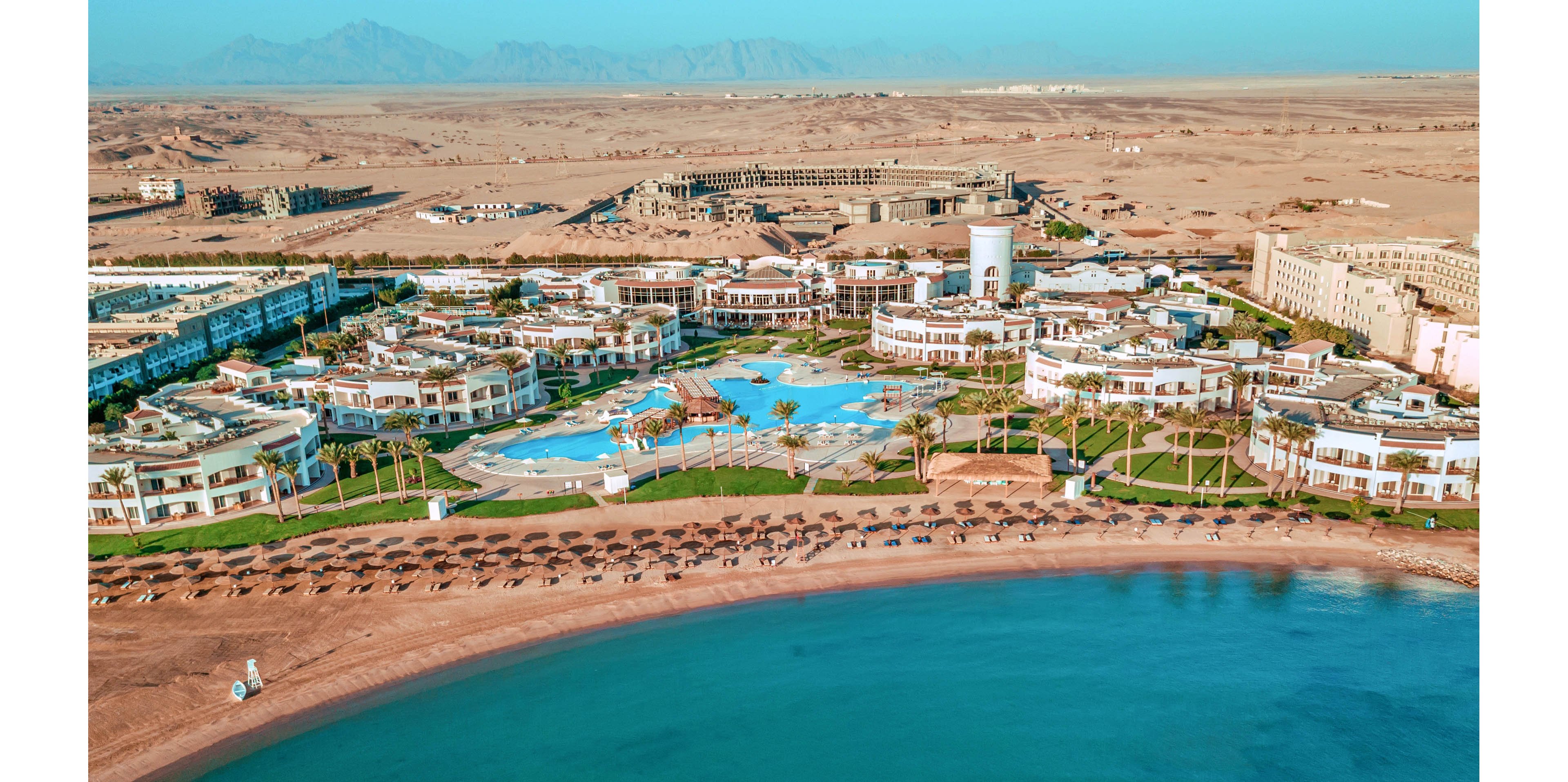 A large coastal resort complex is captured from above, bordered by a calm turquoise sea and a wide sandy beach. Multiple swimming pools, sun loungers, and landscaped green areas are arranged among white low-rise buildings. Rows of beach umbrellas line the shoreline, with a lifeguard or service station near the water. In the background, an expansive desert plain stretches toward distant mountains under a clear blue sky.