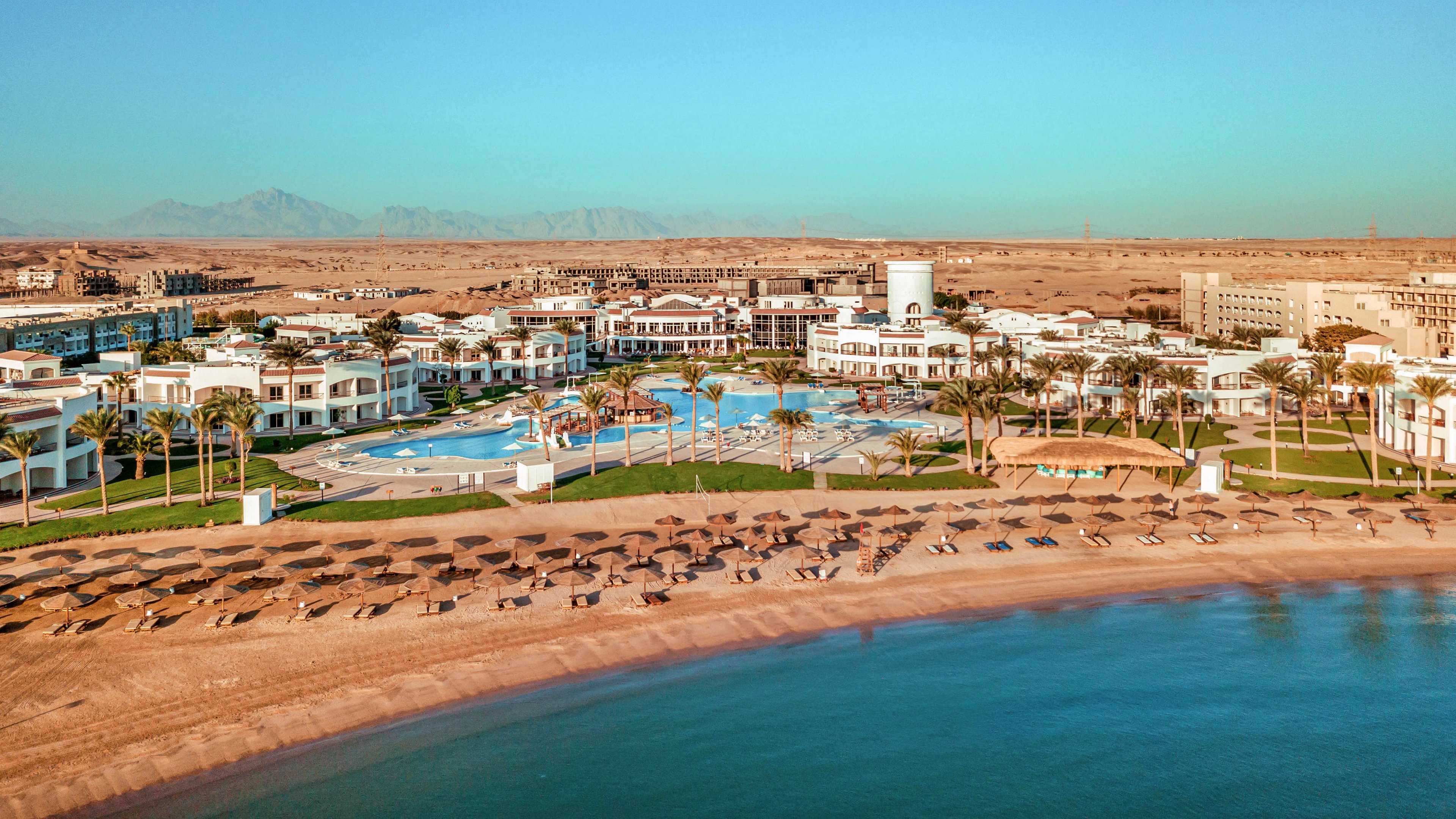 A large seaside resort stretches along a sandy beach with rows of sun loungers and umbrellas facing calm blue water. White low-rise buildings surround expansive swimming pools and palm trees, creating a modern vacation complex. Beyond the resort, a wide desert landscape and distant mountains are visible under a clear blue sky. The scene suggests a tranquil, sunny holiday destination with ample outdoor relaxation areas.