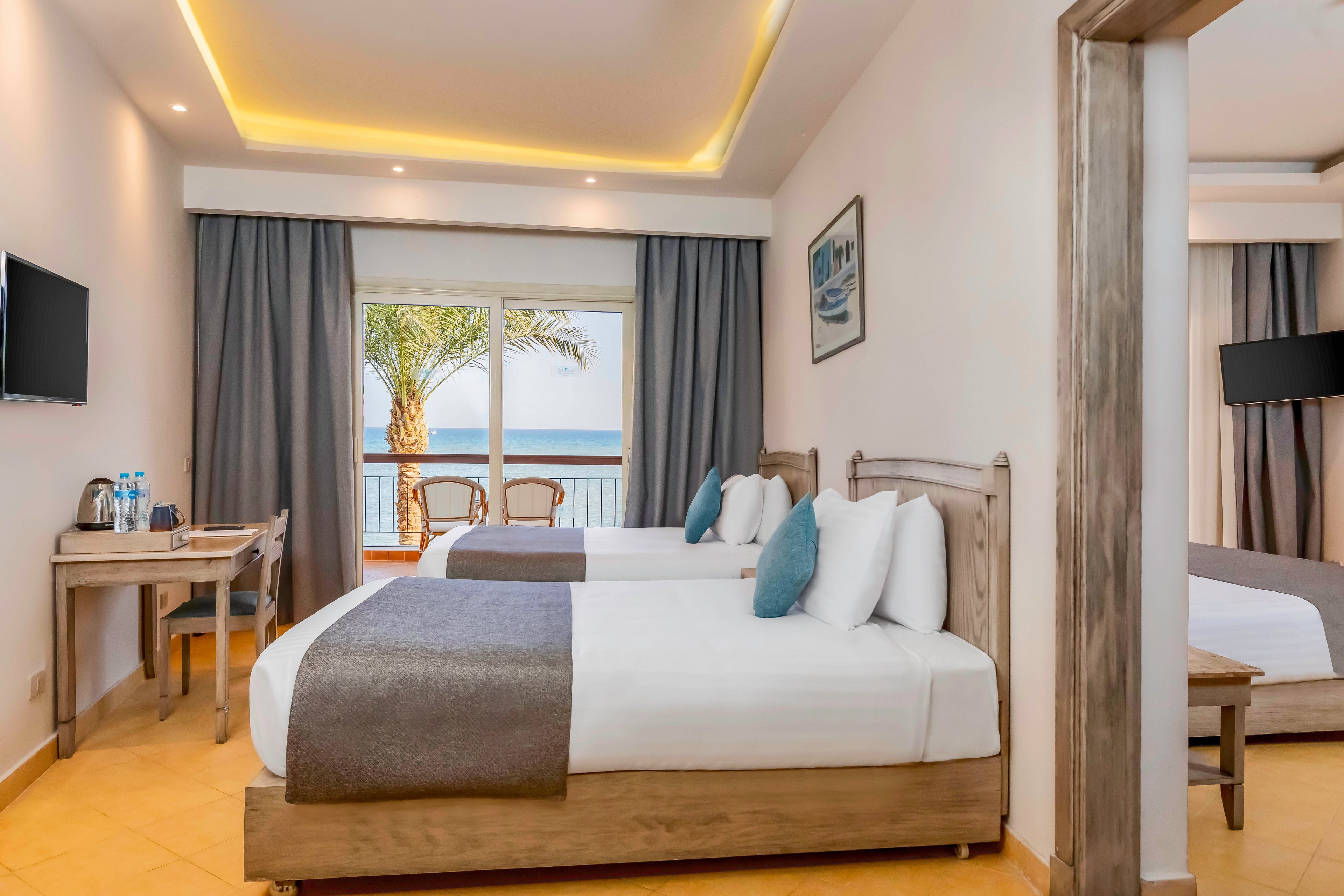 Modern hotel room featuring two neatly made double beds with white linens and gray throws. A balcony with wooden chairs overlooks the ocean and a palm tree through a large glass door. The room includes a wall-mounted television, a small desk with bottled water and a kettle, and neutral decor with blue accent pillows. Soft ceiling lighting and tiled flooring create a clean, inviting atmosphere suitable for vacation or business travel.