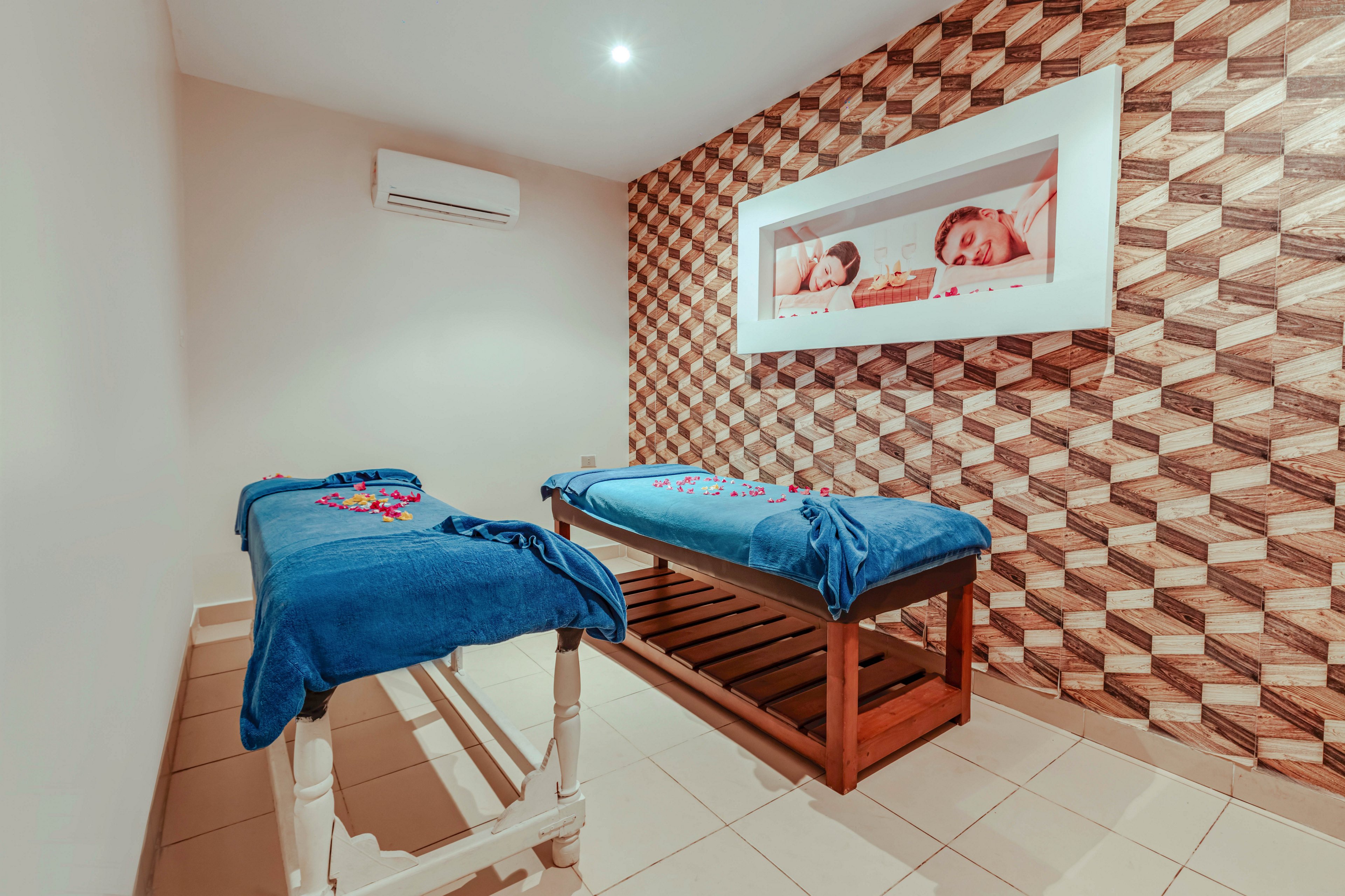 A small spa treatment room features two massage tables covered with blue sheets and scattered flower petals. The space has tiled flooring and a patterned geometric accent wall that adds visual interest. An air conditioning unit is mounted on the plain wall, suggesting a comfortable indoor temperature. A framed photo of two smiling children hangs above one table, contributing a warm and welcoming atmosphere.