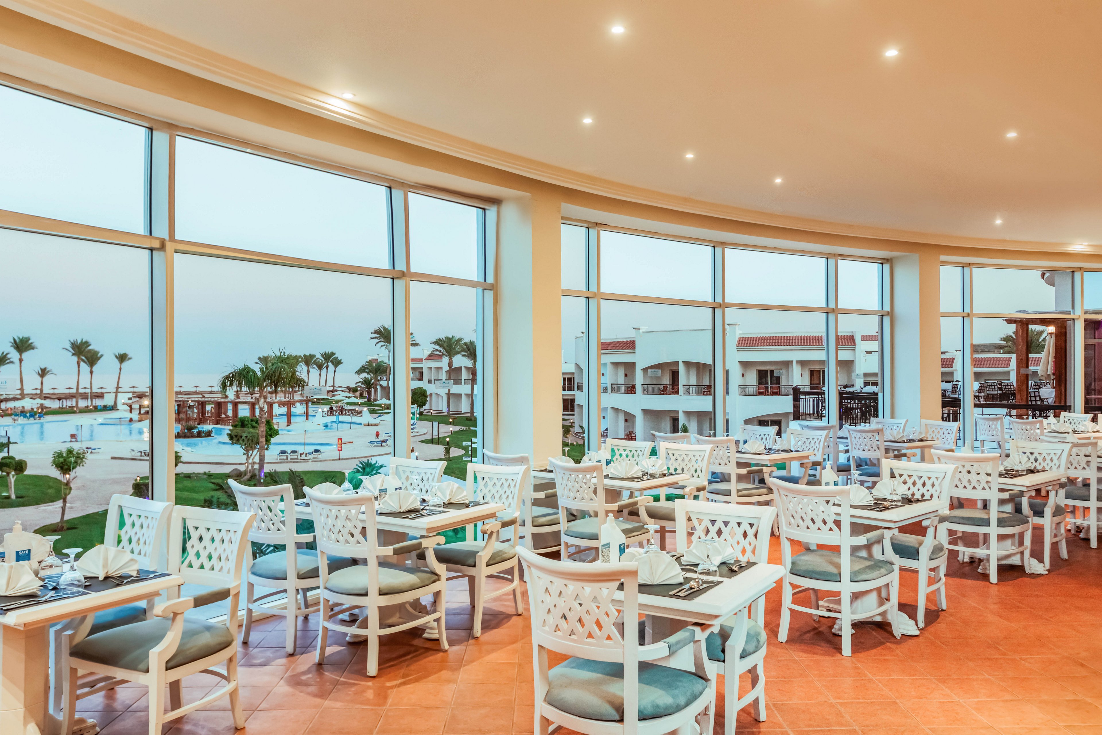 Spacious resort restaurant with white lattice-back chairs and neatly set tables faces large floor-to-ceiling windows. Outside, a swimming pool area with sun loungers and palm trees stretches toward low-rise hotel buildings. Soft indoor lighting and pastel blue cushions create a relaxed, coastal atmosphere. The scene suggests a vacation dining experience with views of the pool and landscaped grounds.