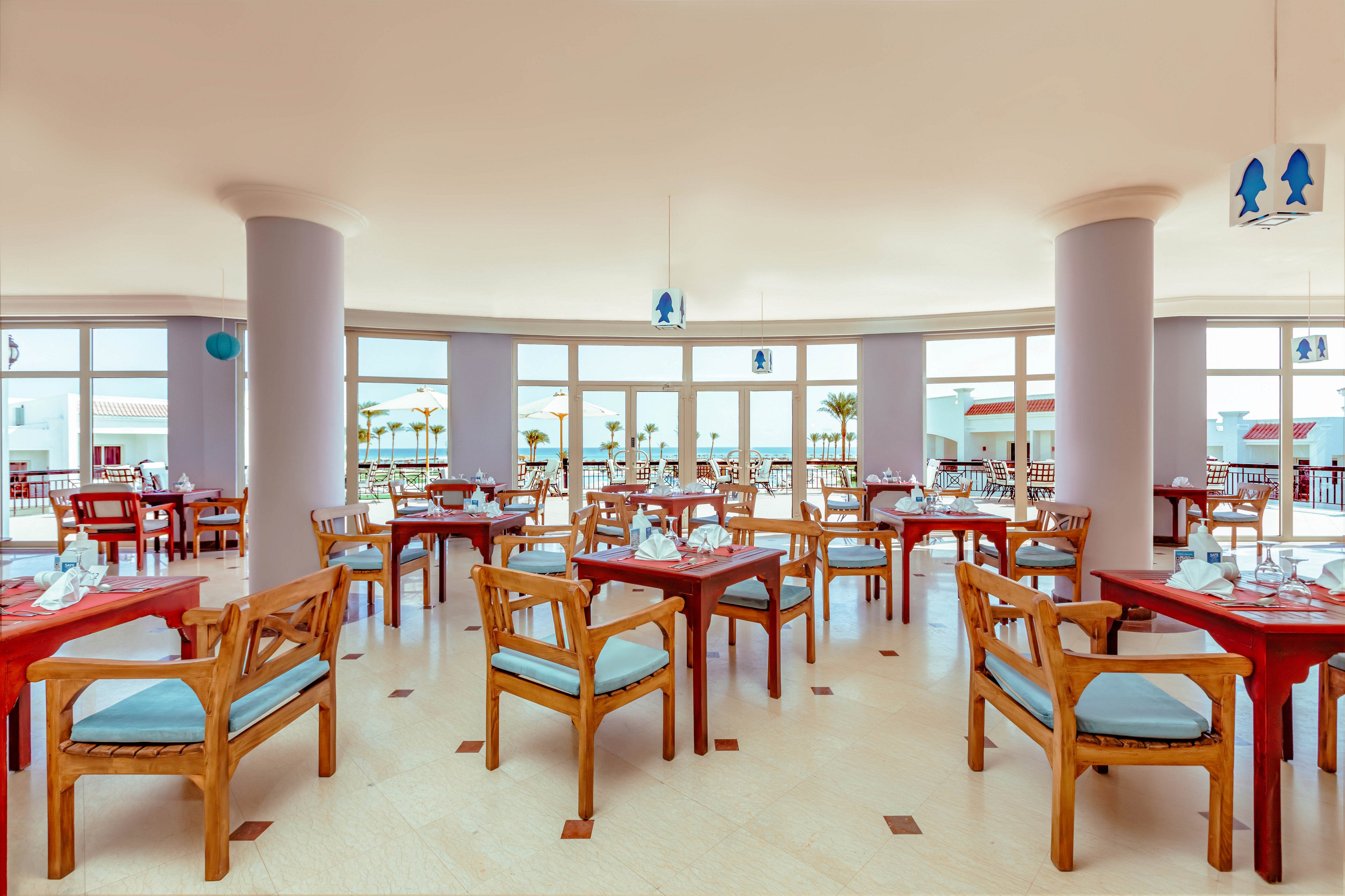 Spacious restaurant dining area with wooden tables and cushioned chairs is shown in warm natural light. Large floor-to-ceiling windows reveal an outdoor terrace and distant sea view. Tables are neatly set with glassware, plates, and folded napkins, suggesting readiness for guests. Soft pastel colors and clean, open layout create a relaxed coastal atmosphere.