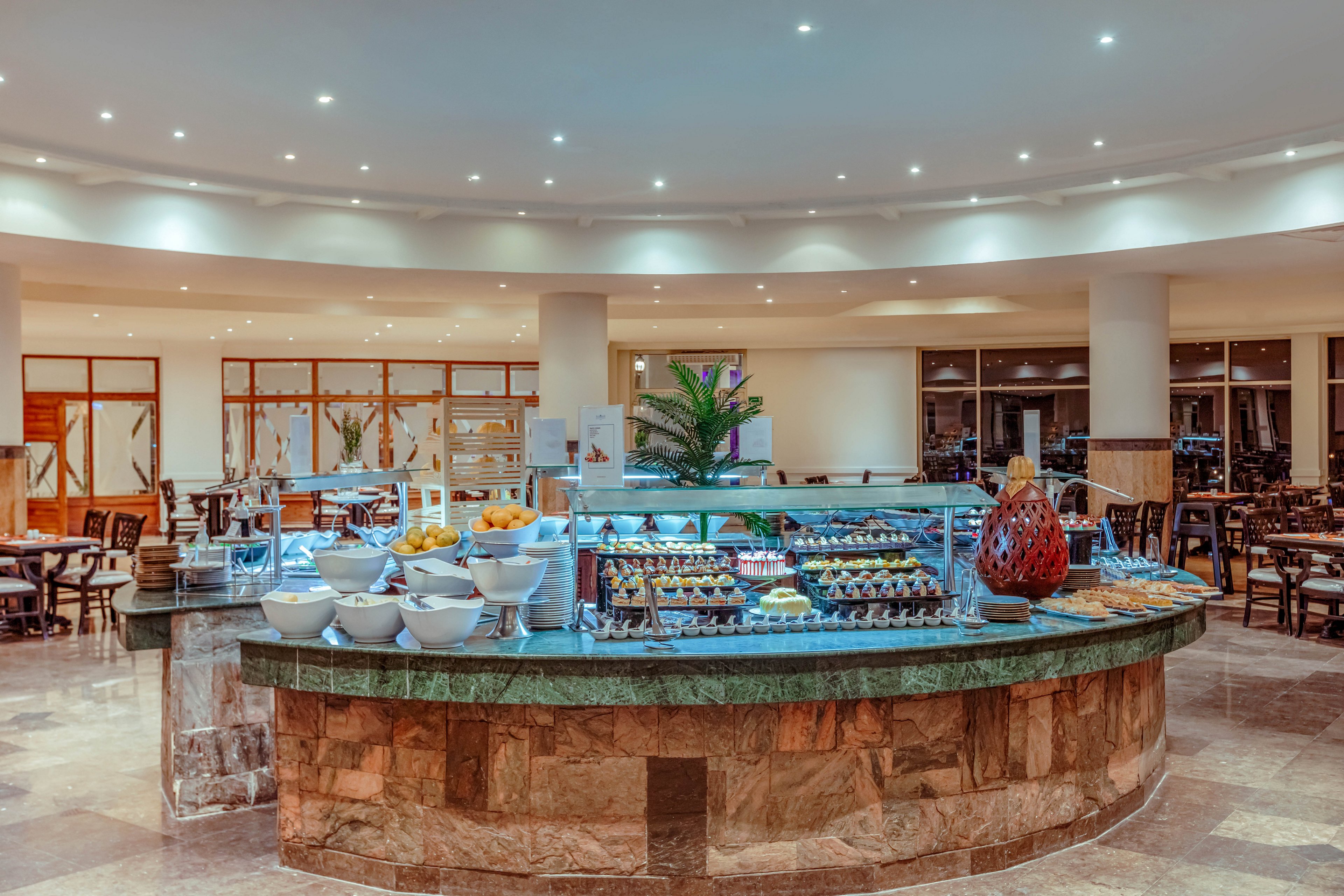 A large circular buffet counter stands in the center of a spacious restaurant interior. The counter displays an assortment of dishes, desserts, and bowls arranged neatly under bright ceiling lights. Stone and green marble surfaces give the area a polished, upscale look. In the background, dining tables and chairs are set for guests, creating a welcoming, modern atmosphere.