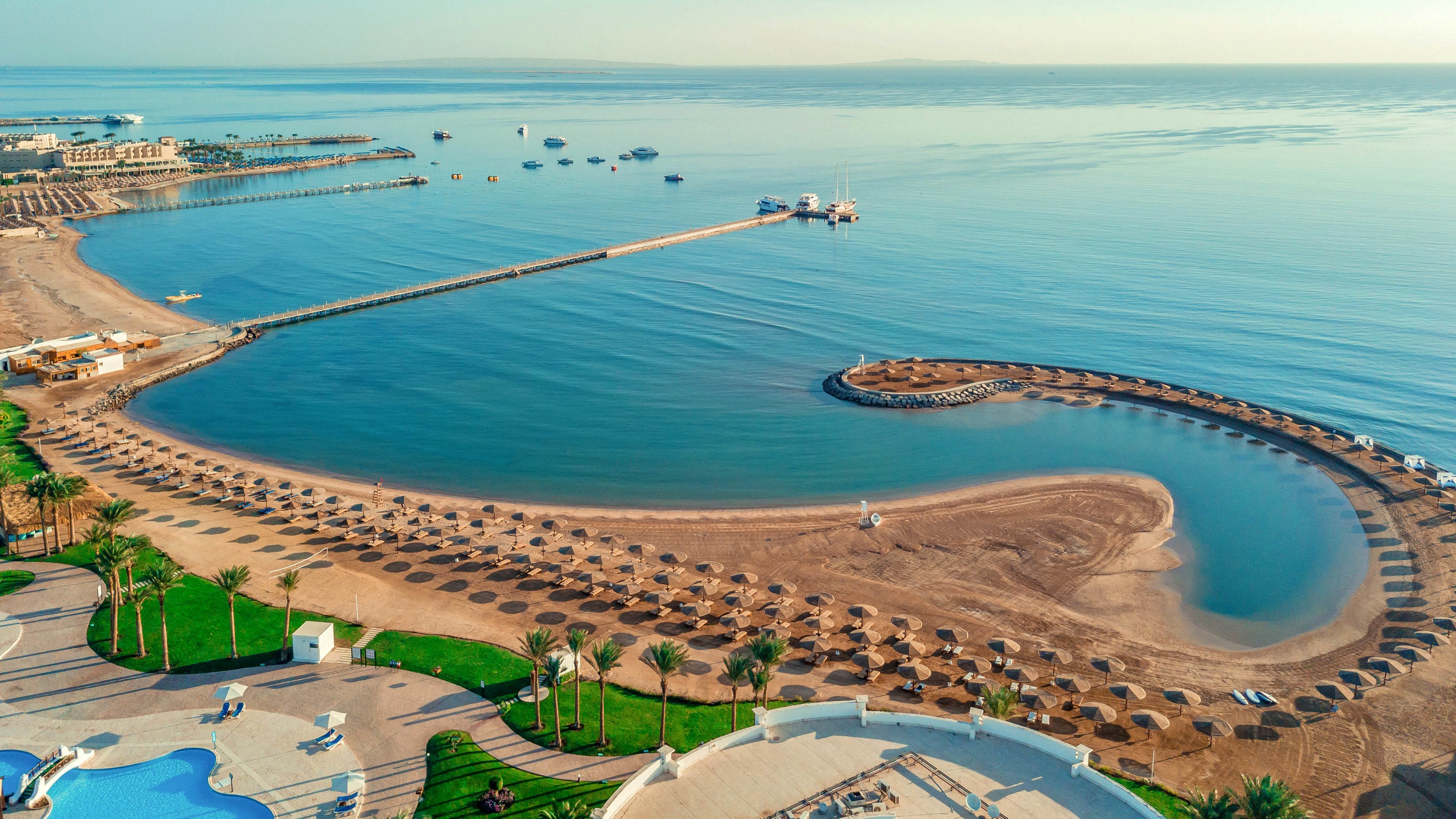 A curved sandy bay with neatly arranged beach umbrellas stretches along a calm, clear sea. A long pier extends into the water, with boats anchored in the distance. Resort buildings, pools, and palm trees frame the shoreline, creating a luxurious holiday setting. The overall scene feels peaceful and inviting, ideal for relaxation and seaside vacations.