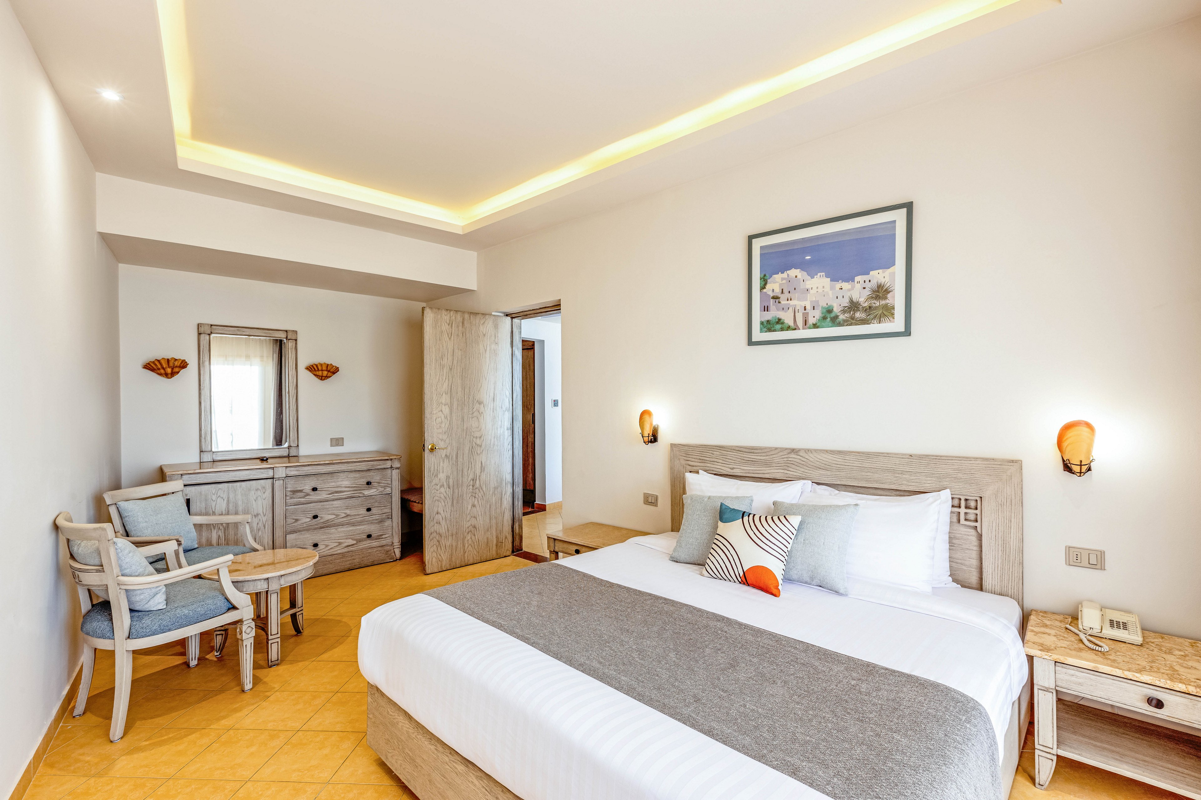 A neatly made double bed with white linens and a gray runner is the focal point of this bright hotel bedroom. The room features light wooden furniture, including bedside tables, a dresser with mirror, and a small table with two chairs. Warm wall sconces and recessed ceiling lighting create a welcoming atmosphere against the neutral walls. A framed artwork above the bed adds a touch of color and character to the minimalist interior.