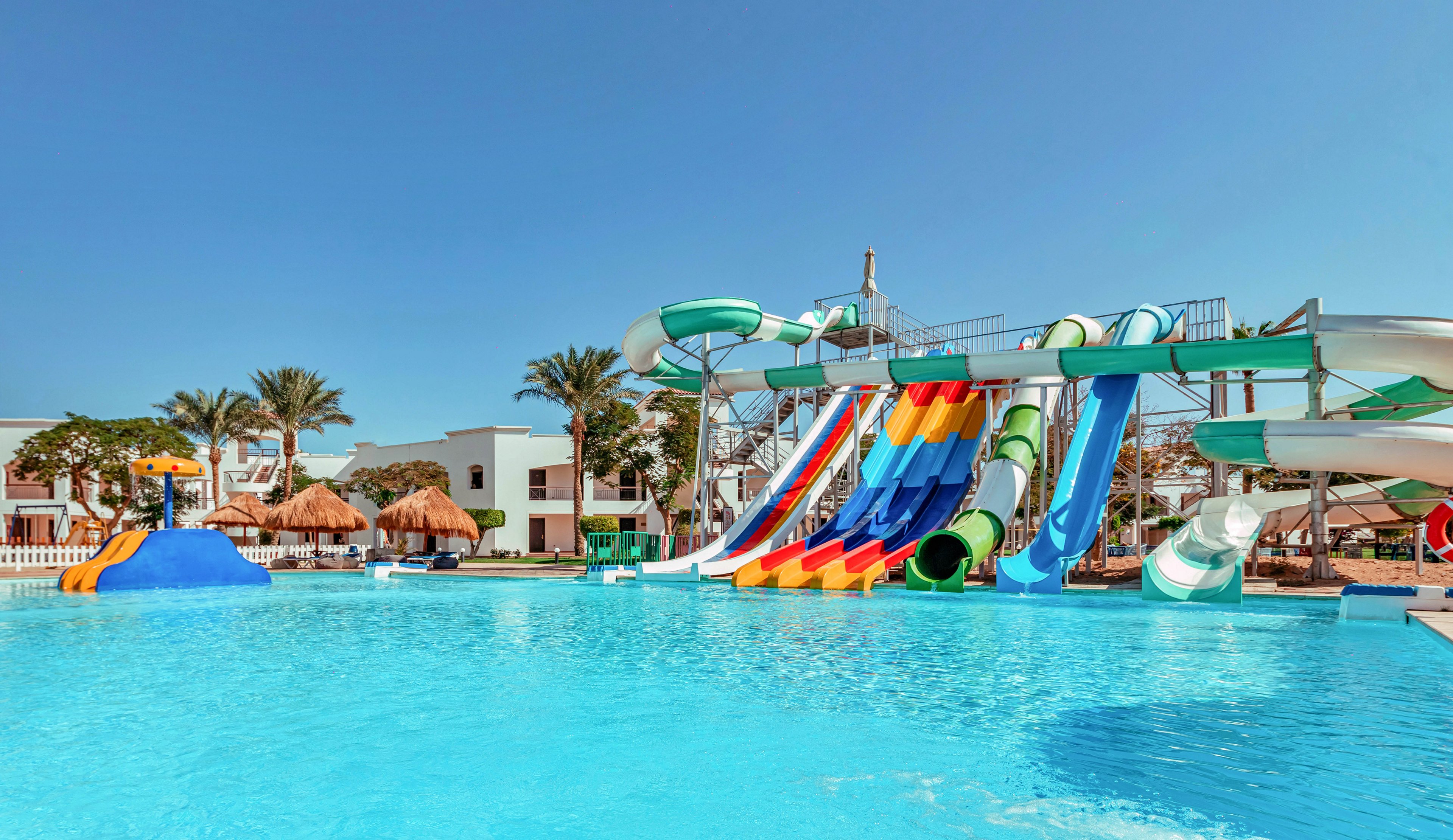 A large outdoor swimming pool is shown in front of a colorful array of water slides and tubes. The setting appears to be a resort with palm trees, thatched umbrellas, and white buildings in the background. Bright blue water reflects the clear sky, emphasizing a sunny, warm climate. The scene suggests leisure, fun, and family-friendly recreation at a vacation destination.