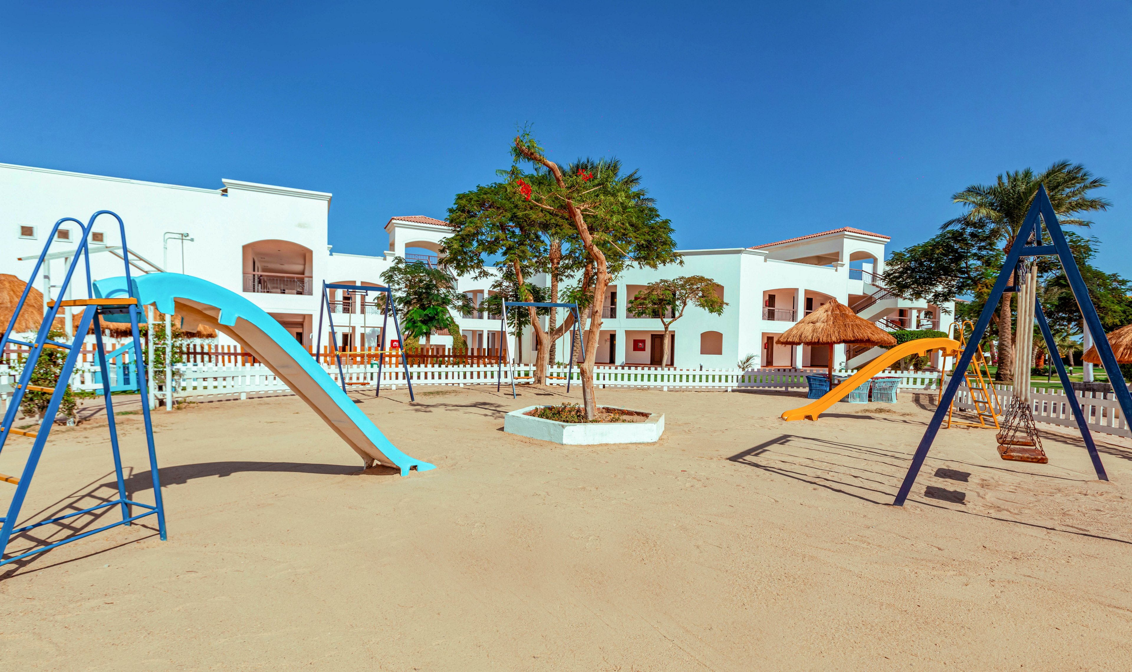 An empty playground with bright, colorful slides stands on a sandy surface under a clear blue sky. In the background, white resort-style buildings with balconies and thatched-roof structures are visible behind a white fence. A few trees add greenery to the otherwise open, sunlit space. The scene suggests a quiet holiday setting ready for children and families.