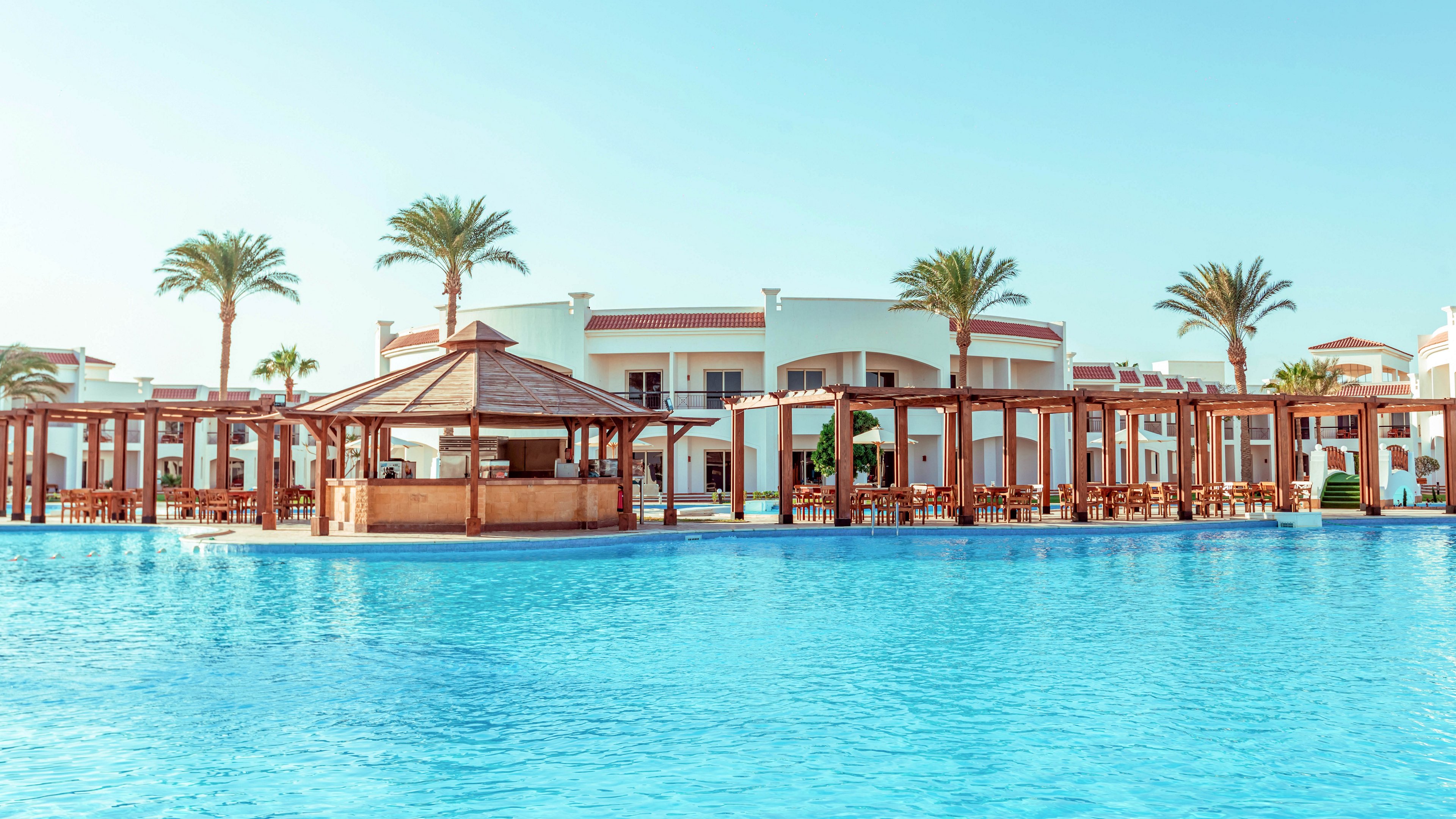 A large turquoise swimming pool stretches across the foreground of a luxury resort. White low-rise buildings with curved facades and red accents line the background, framed by tall palm trees. A wooden poolside bar and shaded pergola seating area sit beside the water, suggesting a relaxing vacation setting. The clear blue sky and warm sunlight create a tranquil, tropical atmosphere.