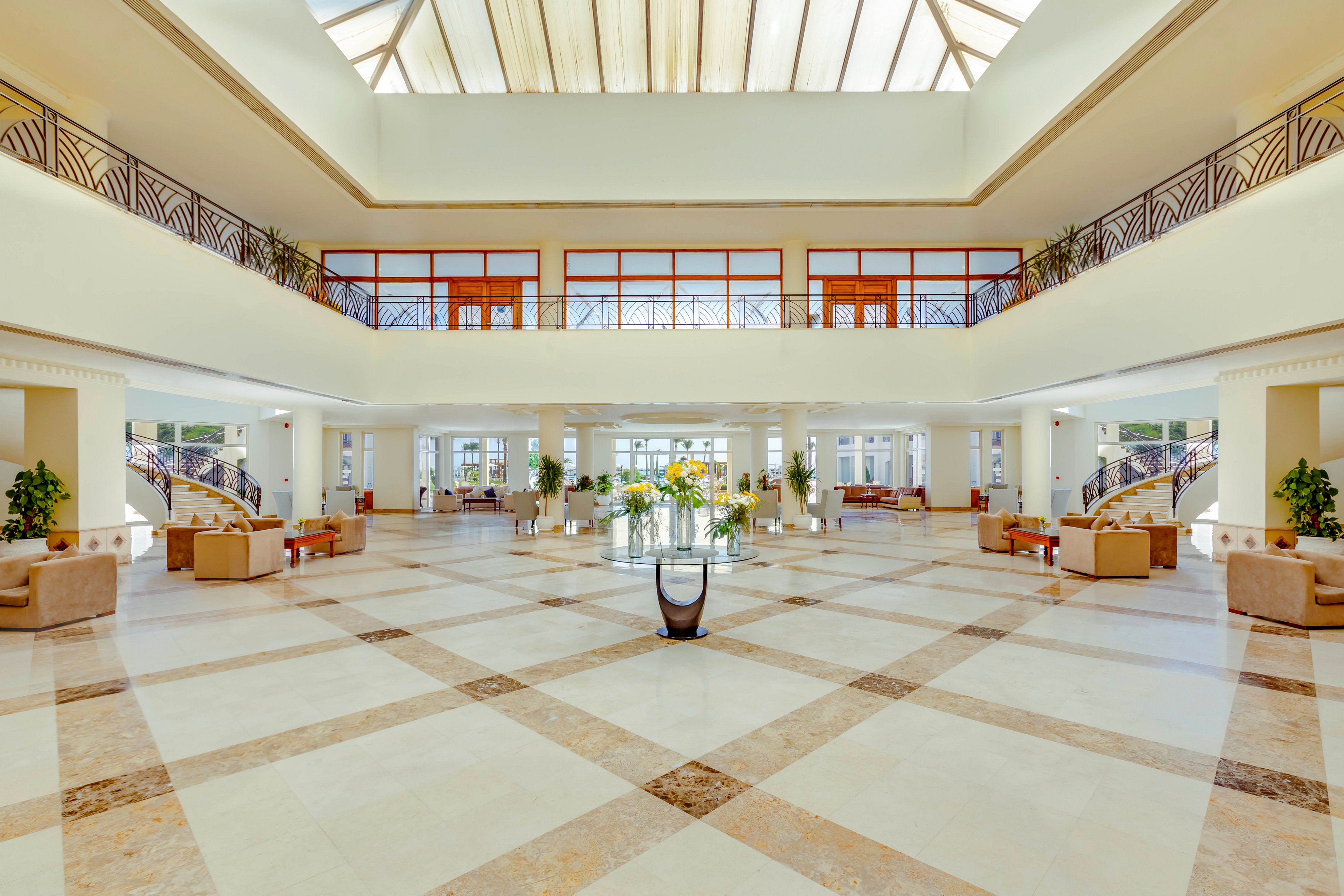 Bright, expansive hotel lobby interior with high ceilings and large skylight. Symmetrical marble floor pattern leads toward glass doors and outdoor view. Comfortable armchairs and sofas are arranged in seating areas along both sides. Decorative plants and a central flower arrangement add color to the neutral, elegant space. Upper-level balconies with railings frame the open atrium design.