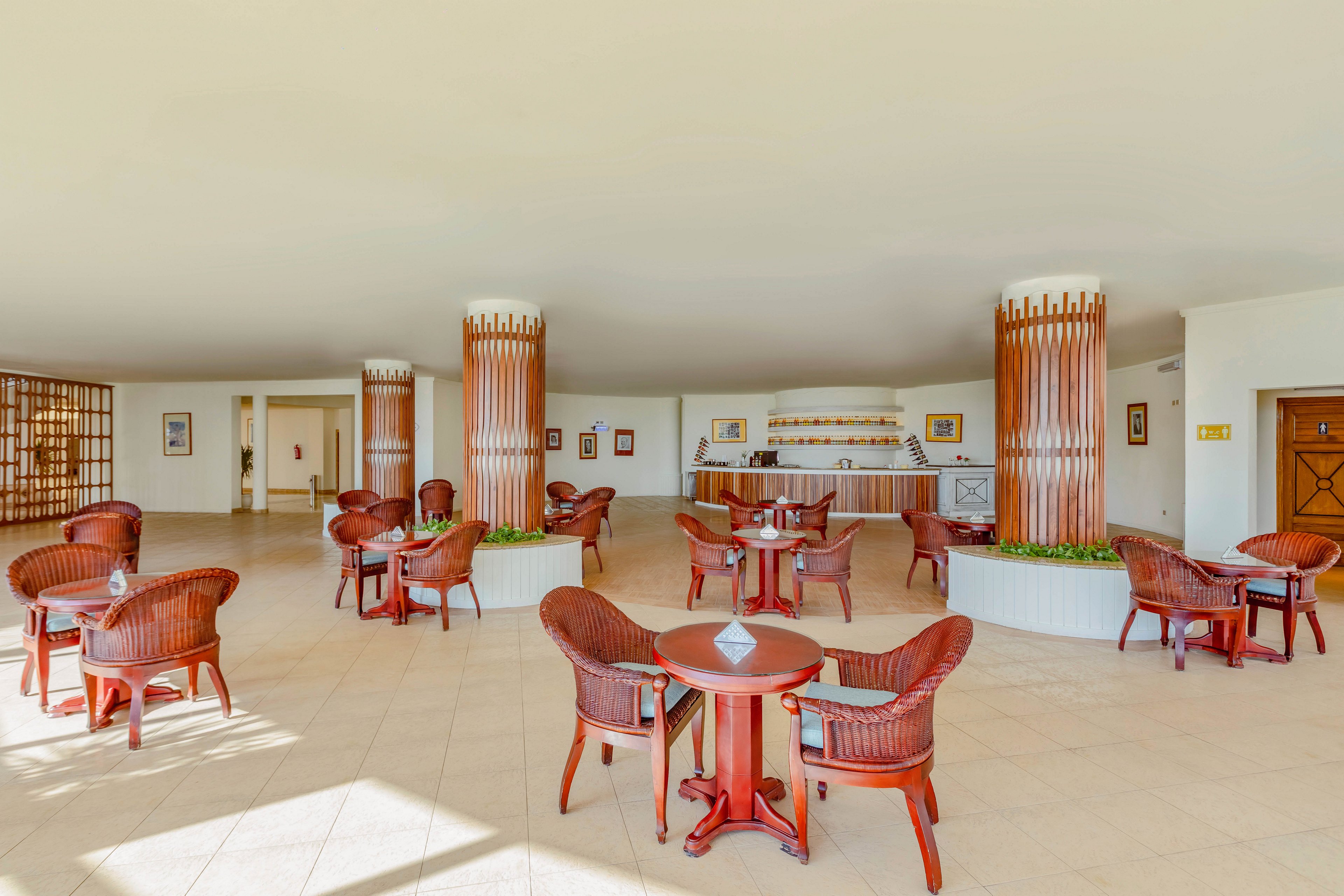 Spacious indoor lounge area features multiple round tables with woven wooden chairs arranged across a tiled floor. Decorative wooden columns frame a small bar counter in the background, stocked with bottles and glassware. The room is brightly lit with neutral walls, framed artwork, and doors leading to adjacent spaces. Overall, the setting suggests a relaxed hotel or resort environment designed for casual socializing and refreshments.