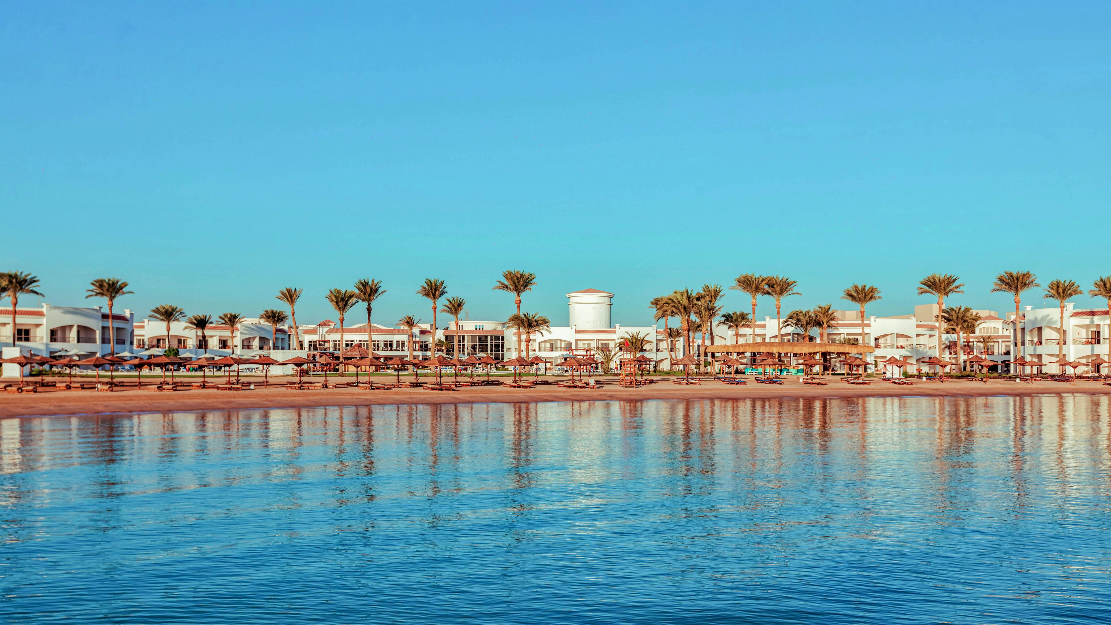 A coastal resort lined with tall palm trees stretches along a sandy shoreline. Low white buildings and beach umbrellas are visible behind the palms, suggesting a popular vacation destination. The calm sea in the foreground reflects the warm colors of the sky, creating a tranquil atmosphere. The clear blue sky and golden light evoke a relaxed, tropical holiday mood.