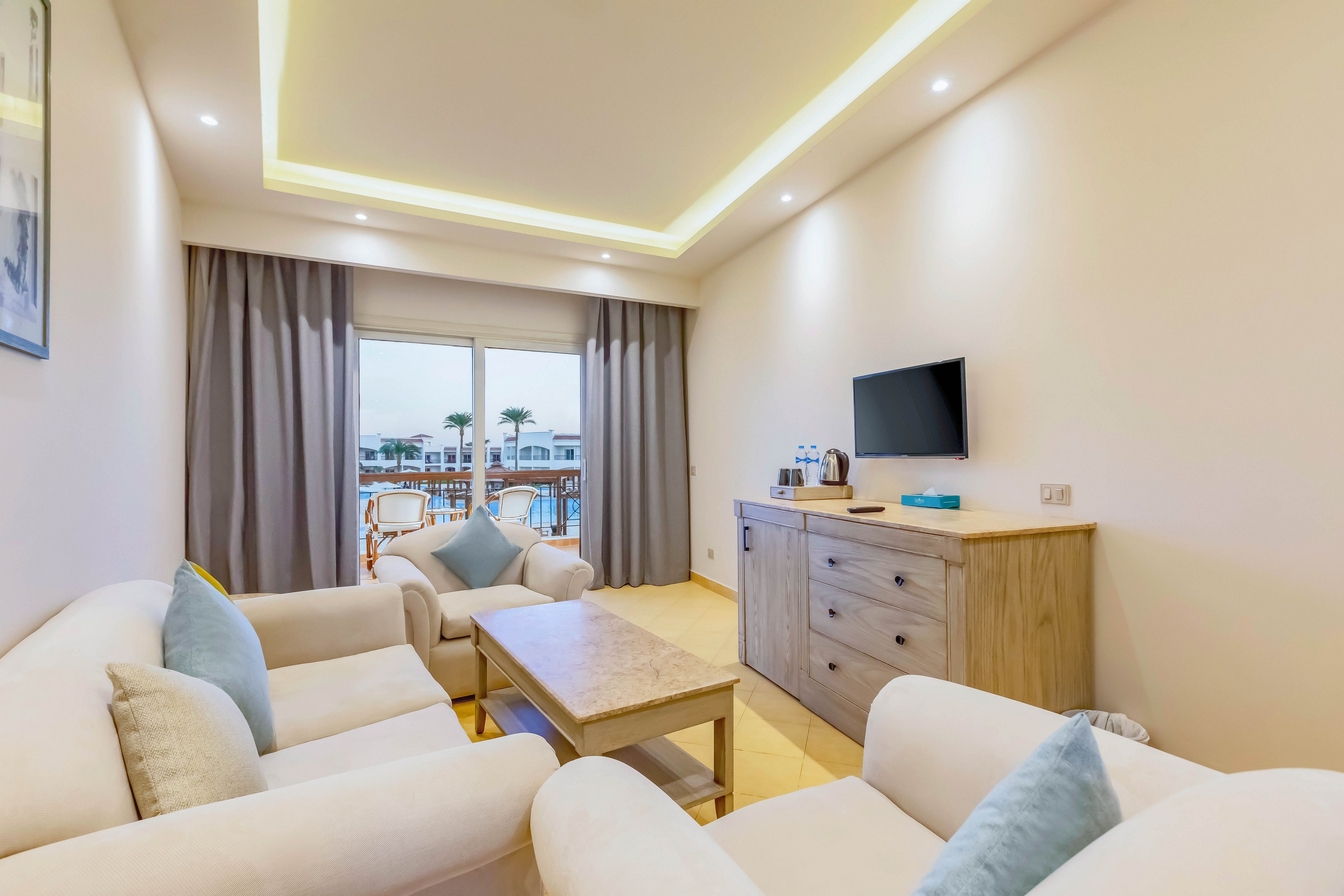 Contemporary hotel suite living room with cream sofas and a wooden coffee table is shown in soft, warm lighting. A flat-screen TV is mounted above a light wood dresser holding a kettle and cups. Large sliding glass doors with gray curtains open to a balcony overlooking a pool and palm trees. The neutral color palette with light blue cushions creates a calm, inviting atmosphere.