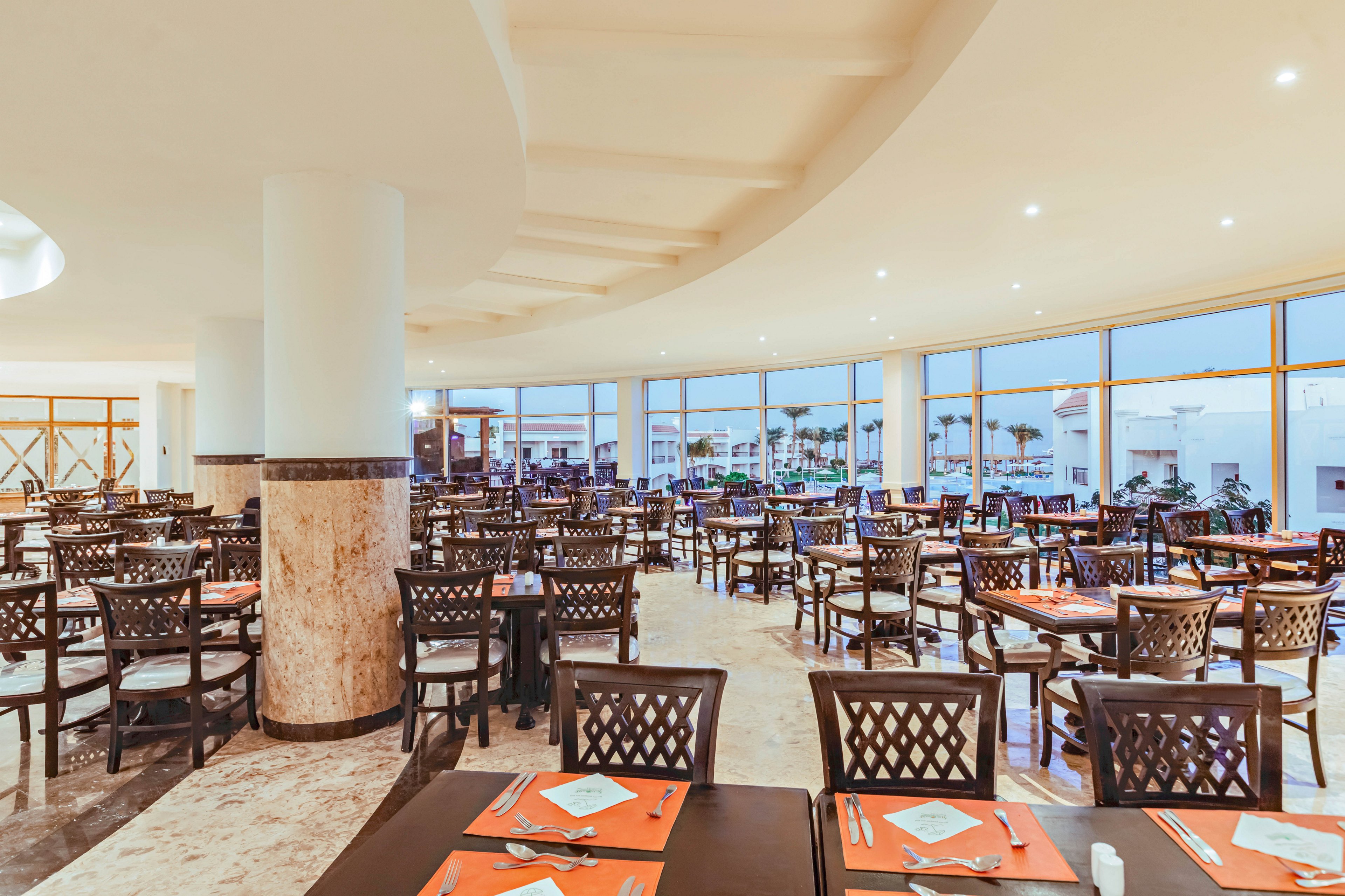 A large, bright restaurant dining area is filled with rows of dark wooden tables and chairs. Each table is neatly set with orange placemats, folded napkins, and cutlery, creating a uniform look. Floor-to-ceiling windows line one side of the room, revealing an exterior view with palm trees and buildings. Warm interior lighting and polished floors contribute to a clean, welcoming atmosphere suitable for group dining.