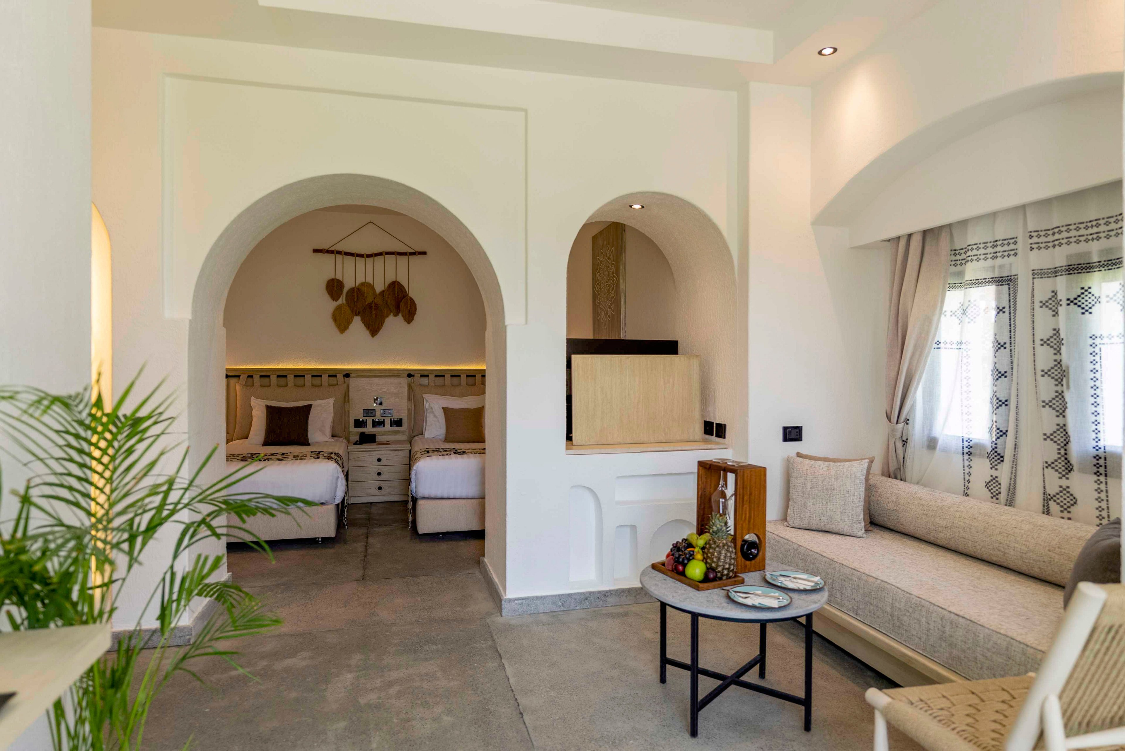 A spacious, light-filled hotel room features an arched alcove with two neatly made twin beds. The interior showcases white walls, natural wood accents, and woven decor elements for a Mediterranean-inspired look. In the foreground, a cozy seating area with a sofa, chair, and small round table displays fruit and decorative items. Large windows with patterned curtains and indoor plants add warmth and a relaxed vacation atmosphere.