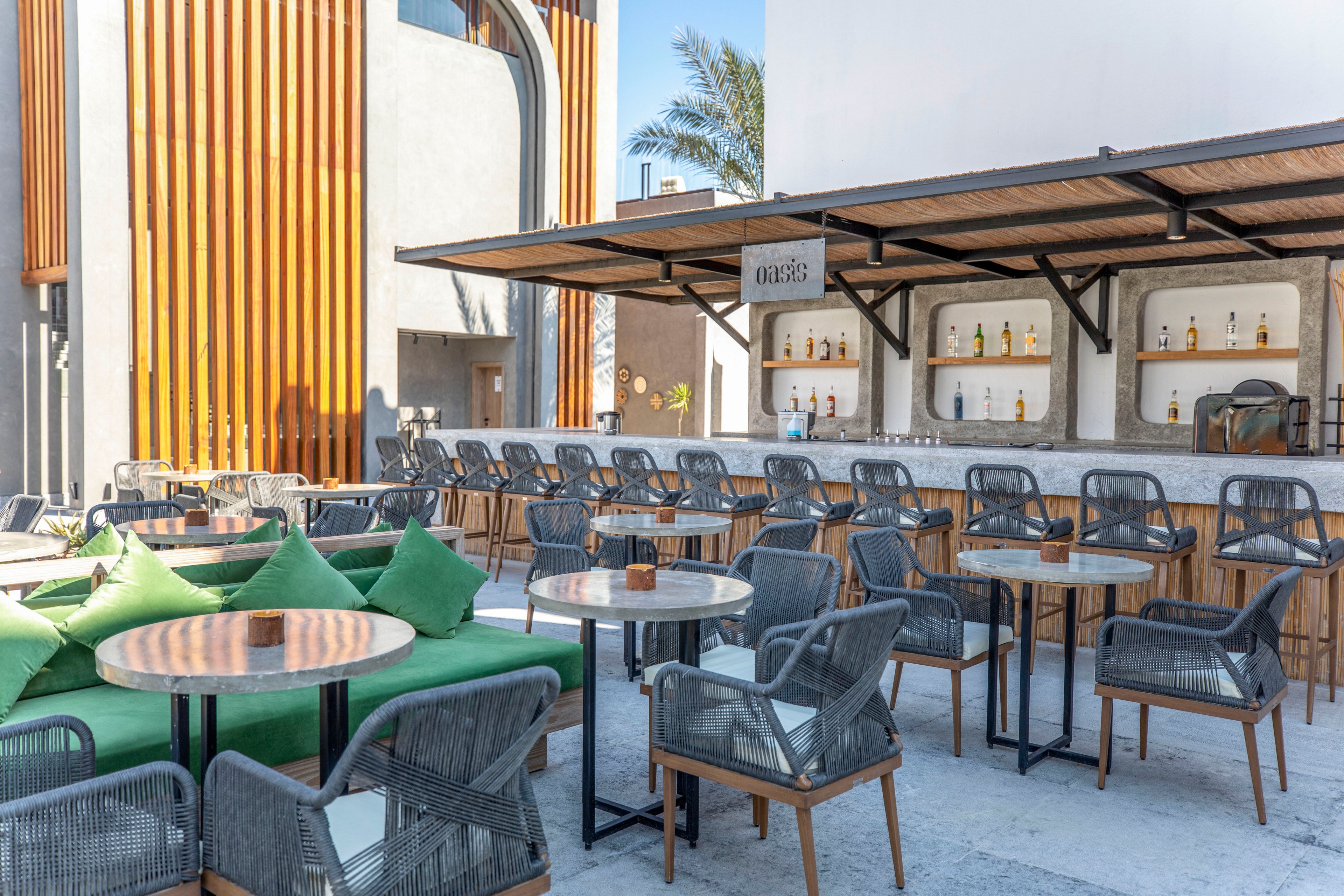An open-air bar features neatly arranged tables and woven chairs in a contemporary setting. The bar counter displays multiple bottles on backlit shelves, suggesting a relaxed drink service area. Green cushions add a pop of color to the neutral-toned furniture and concrete floor. Vertical wooden slats, a white wall, and a palm tree in the background create a warm, modern architectural atmosphere.