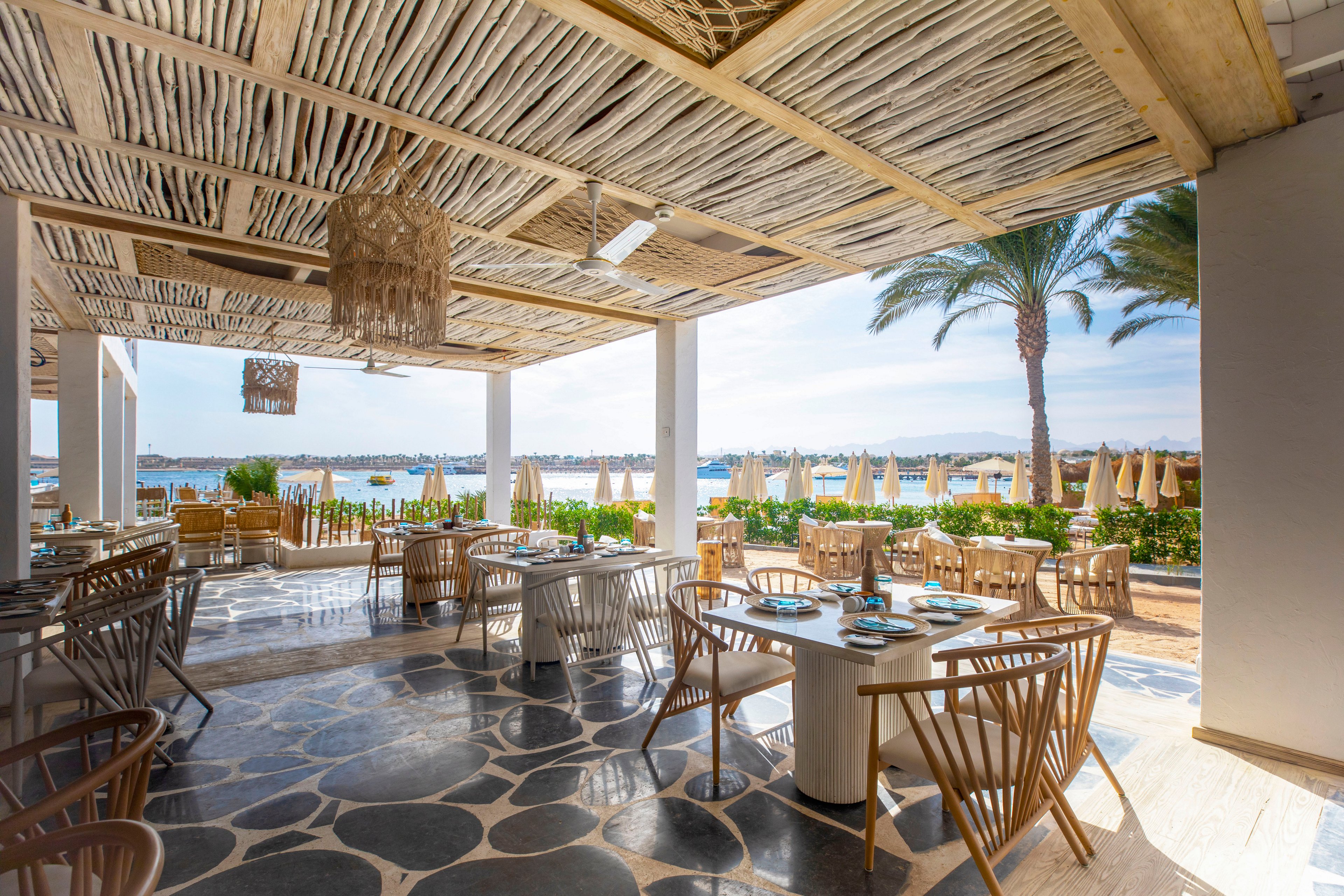 An open-air restaurant with wooden furniture overlooks a sunny seaside landscape. The dining area is shaded by a rustic wooden pergola with woven details and hanging light fixtures. Tables are neatly set, and the floor features a decorative stone pattern. In the background, palm trees, sun loungers, and umbrellas line the waterfront under a clear blue sky.