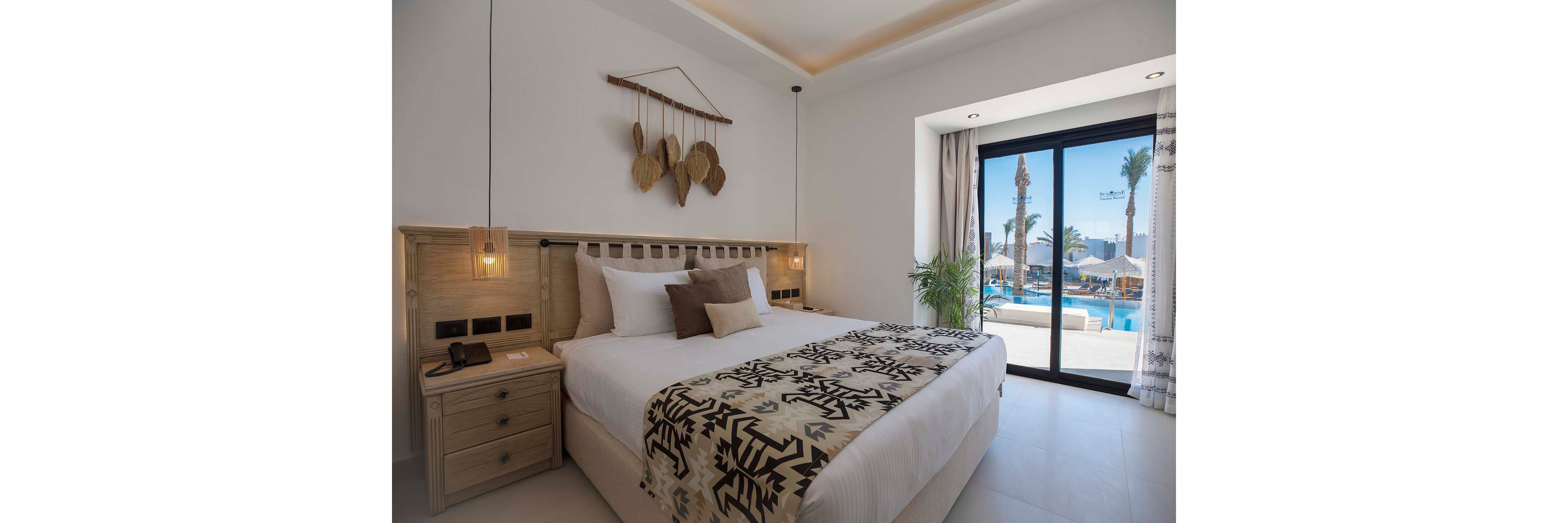 A contemporary hotel bedroom features a large neatly made bed with white linens and patterned throw. Natural wood furniture, including a bedside table with a telephone and hanging pendant lights, adds warmth to the minimalist decor. A sliding glass door opens to a balcony overlooking a bright outdoor pool area with palm trees and sun loungers. Soft neutral tones and natural light create a calm, inviting atmosphere.