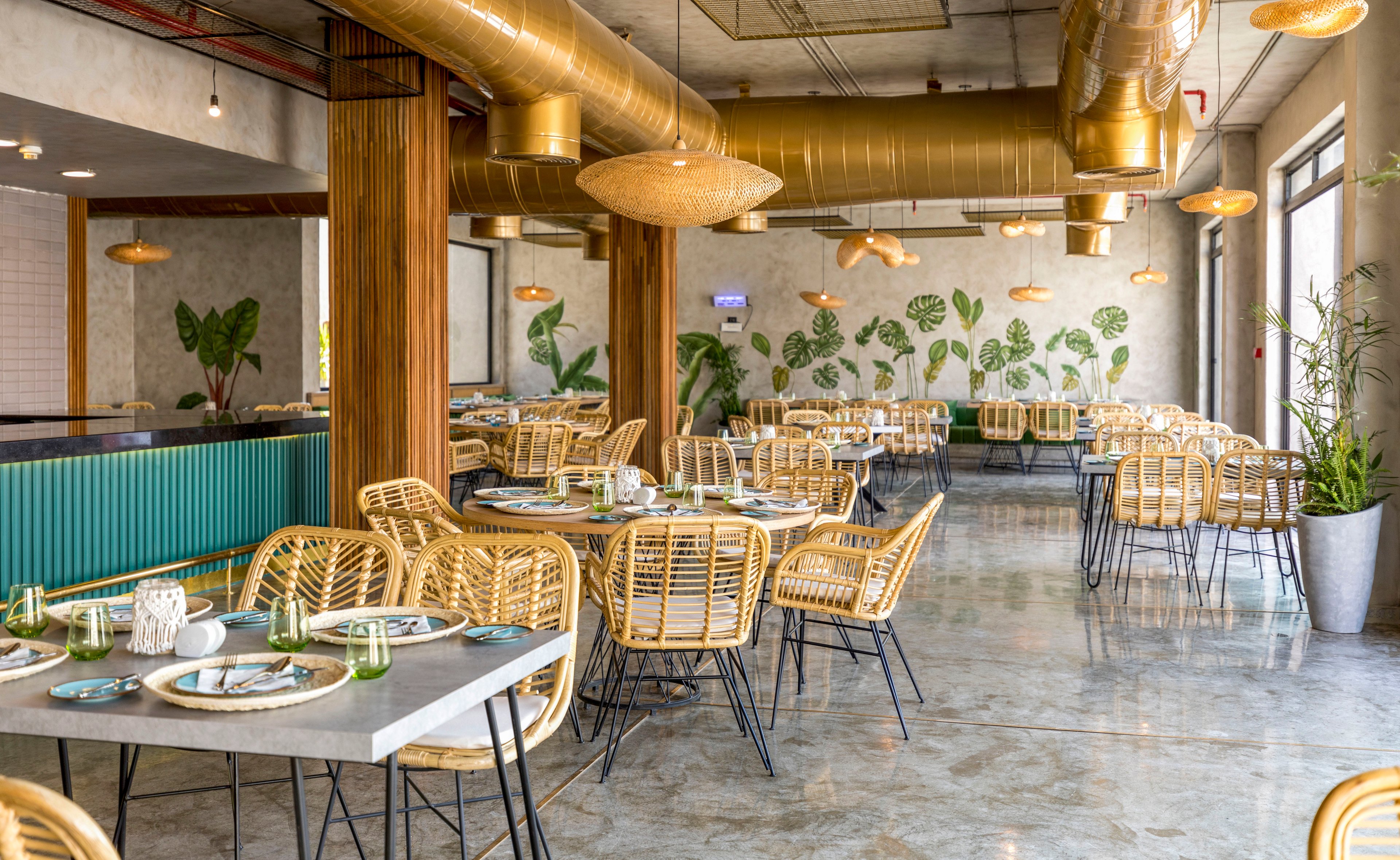 A contemporary restaurant interior features numerous rattan chairs and neatly set tables in a bright, open space. Large windows allow natural light to reflect off the polished concrete floor, highlighting green accents and plants throughout the room. The back wall is decorated with leafy botanical artwork, while gold-colored ceiling ducts and pendant lights add a stylish industrial touch. No people are present, emphasizing the clean, organized dining setup and relaxed tropical atmosphere.