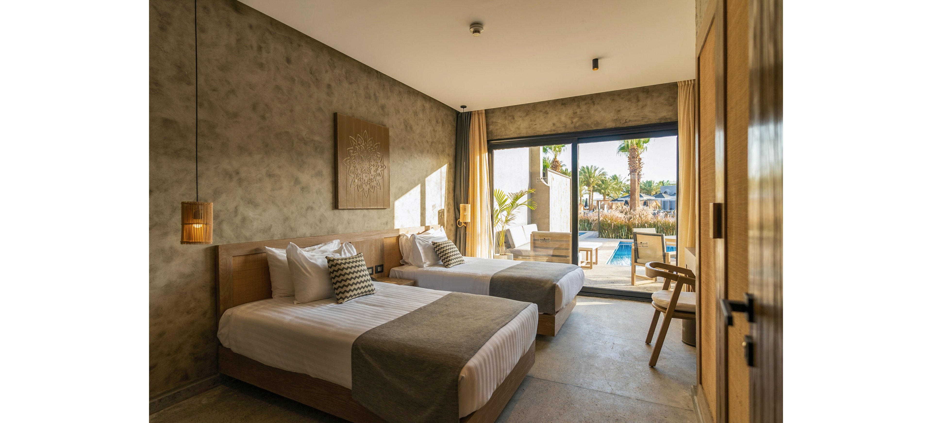 A contemporary hotel room features two single beds with neutral bedding and patterned accent pillows. Warm wooden furniture and textured walls create a minimalist, cozy atmosphere. Large floor-to-ceiling glass doors open onto a private terrace with lounge chairs and a view of a swimming pool and palm trees. Natural light fills the space, highlighting the relaxed resort setting.
